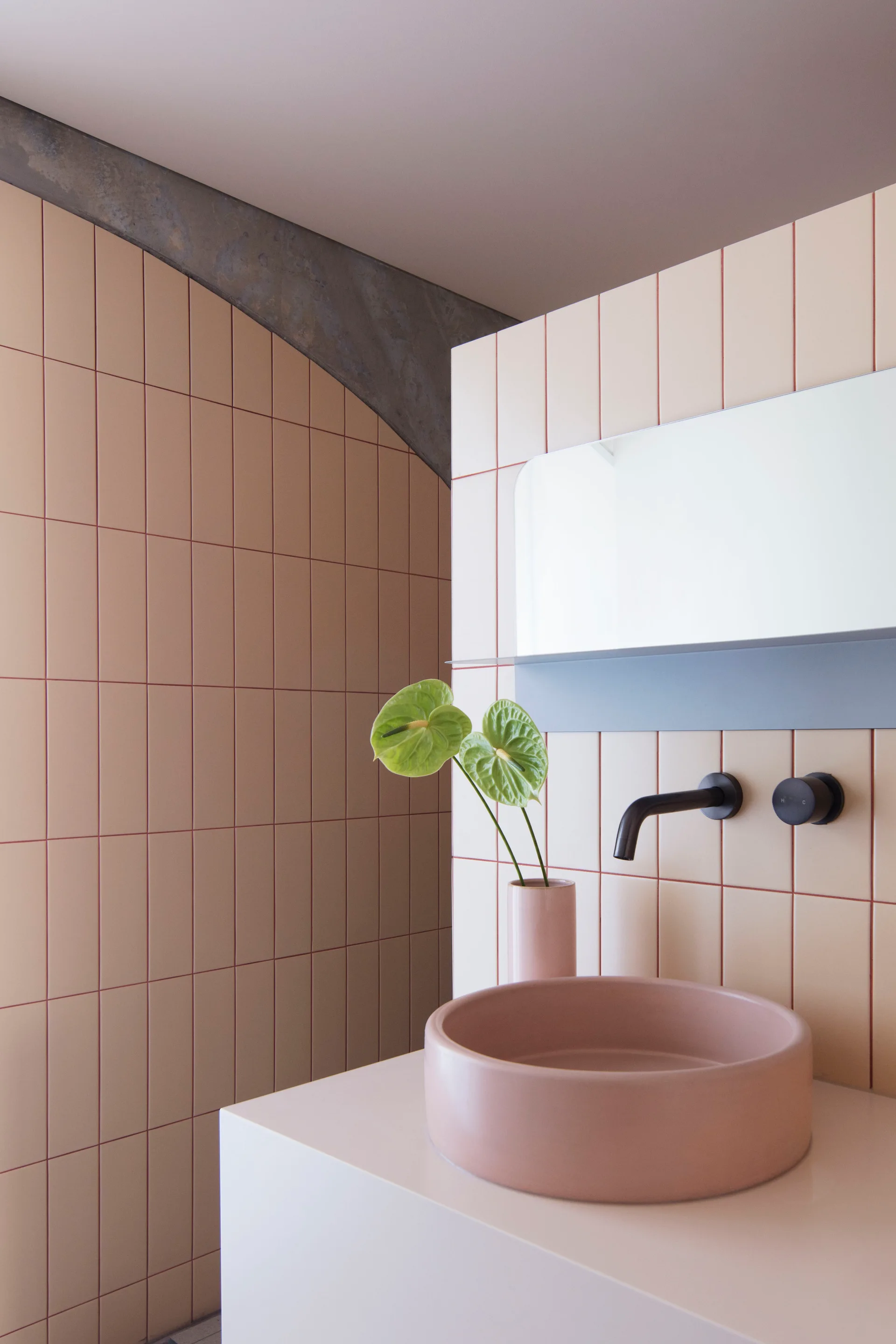 A round pink vessel sink rests on a beige counter against a vertically stacked tiled wall with a wall-mounted matte black faucet.