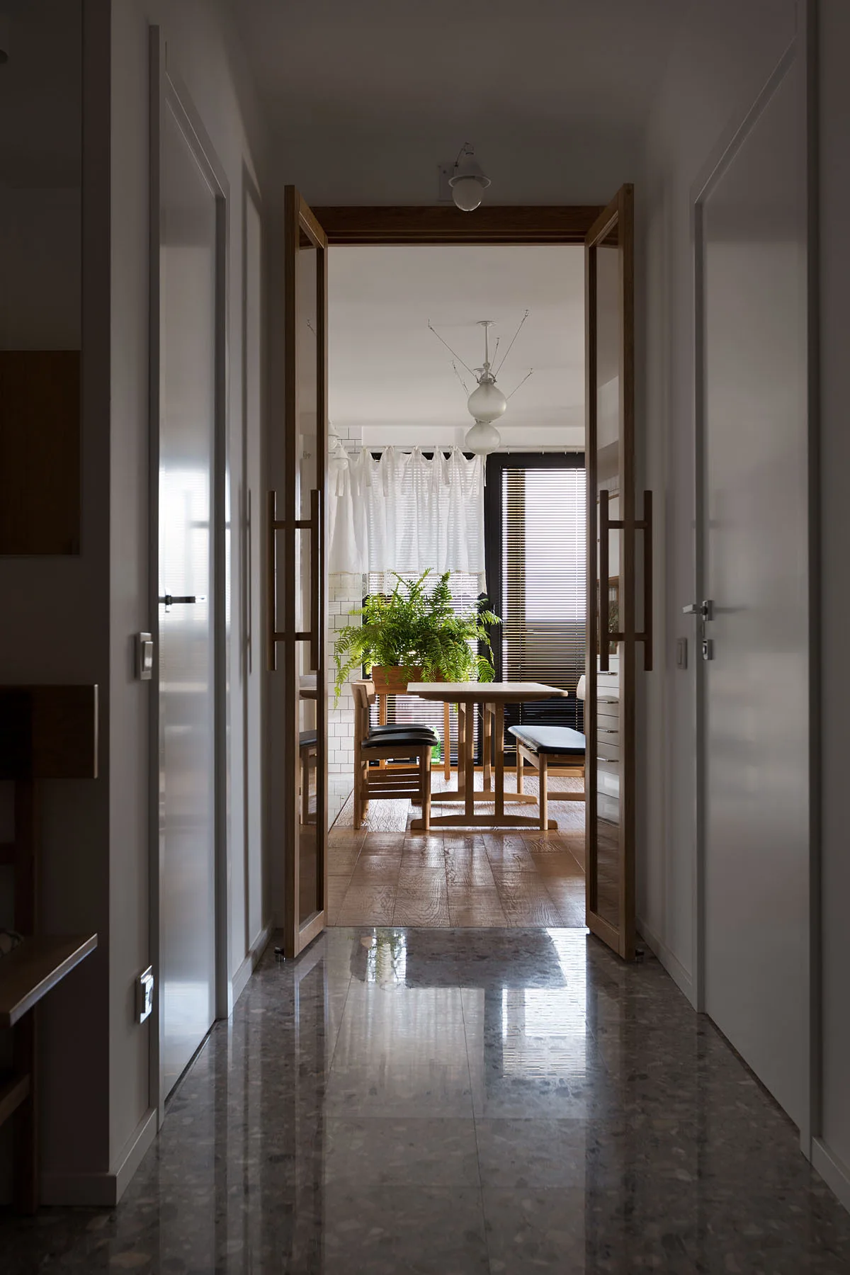A view from a glossy terrazzo-tiled hallway through open wood-framed glass double doors into a sunlit dining area with wooden furniture.
