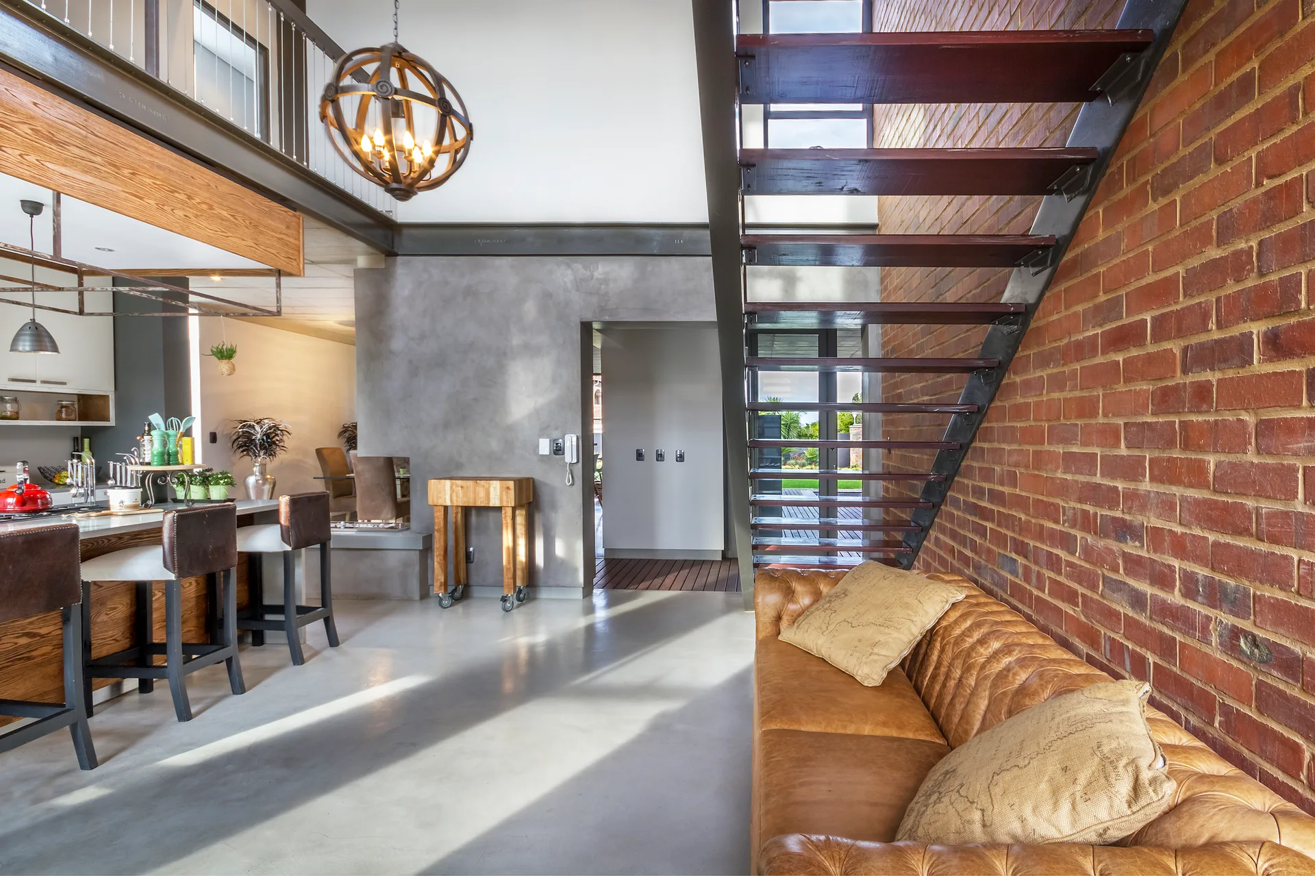 An industrial living area featuring a tan leather Chesterfield sofa against an exposed brick wall next to an open staircase with wooden treads.