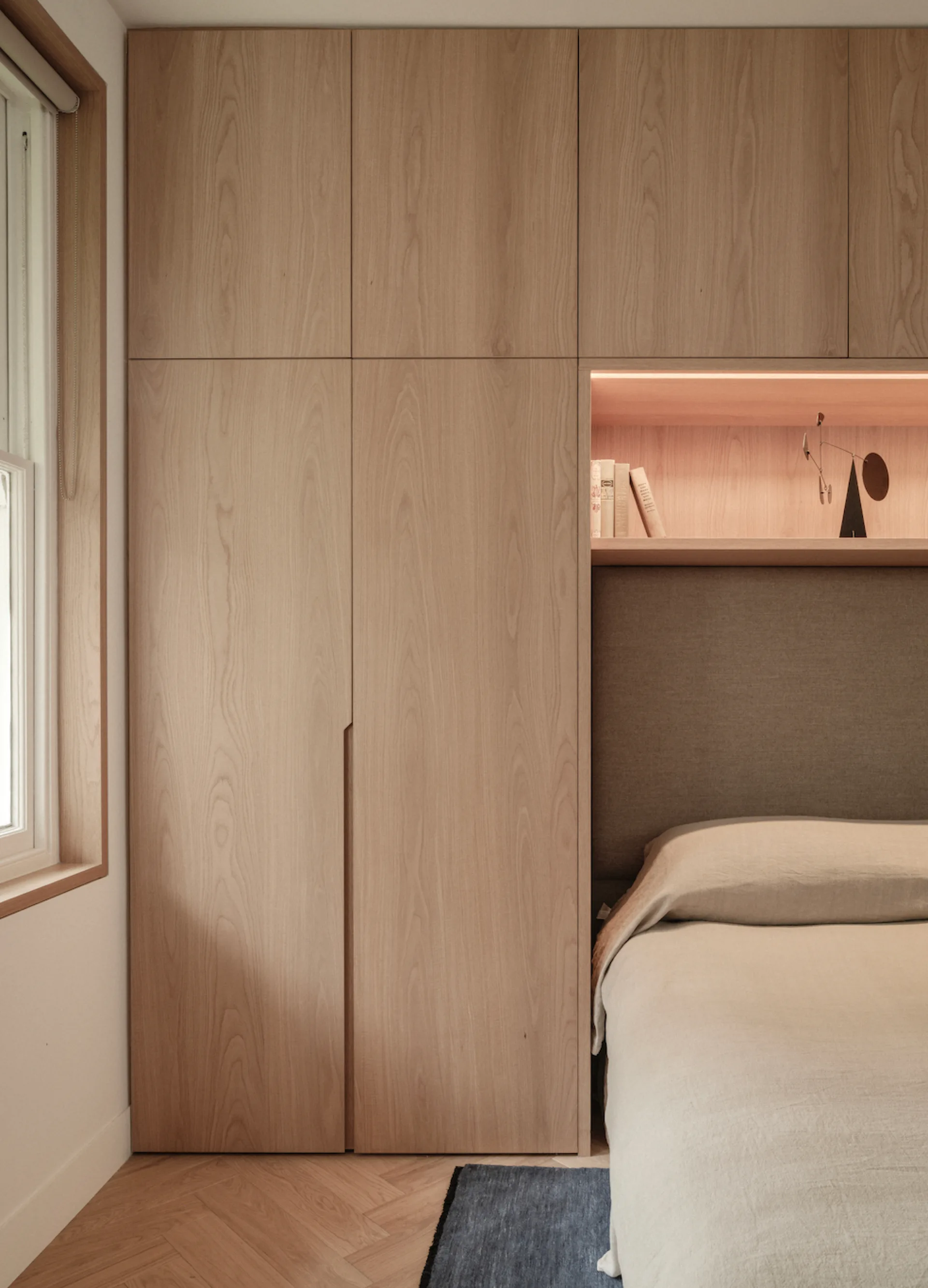 A modern bedroom features seamless light wood cabinetry, a recessed illuminated display shelf, and a bed with a fabric headboard.