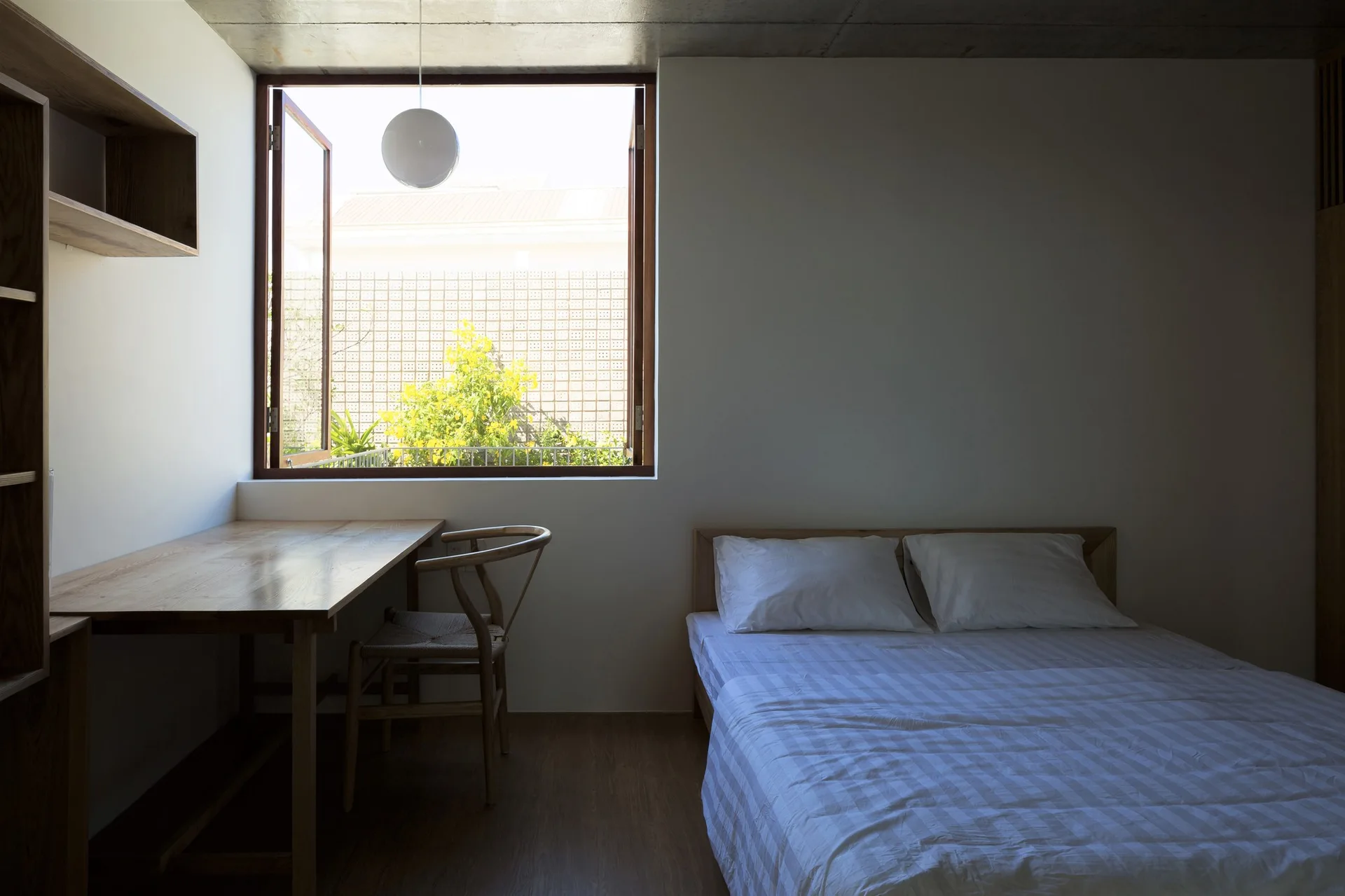 A minimalist bedroom featuring a wooden bed, a study desk with a wishbone chair, and an open window beneath an exposed concrete ceiling.
