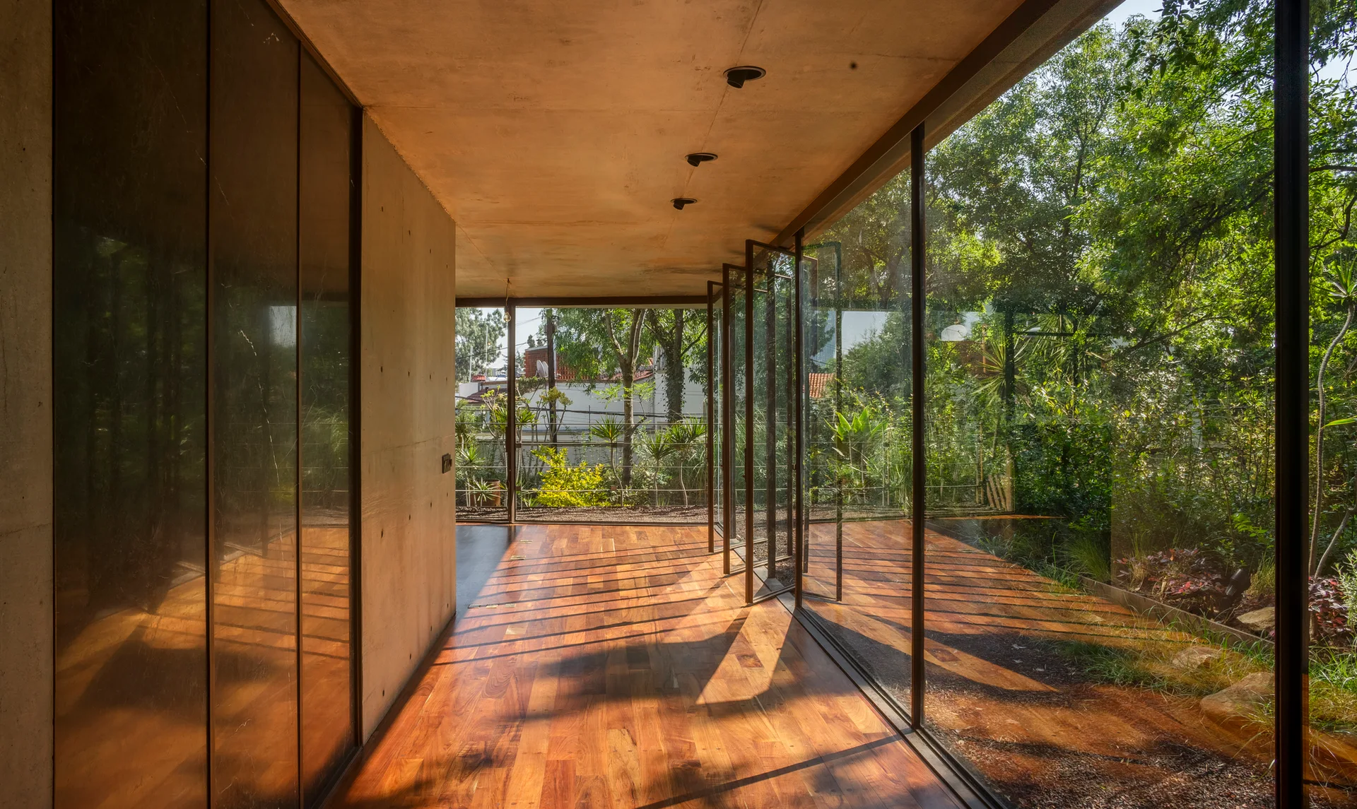 A minimalist corridor features polished wood flooring, a smooth concrete wall, and pivoting glass doors opening to a lush garden.