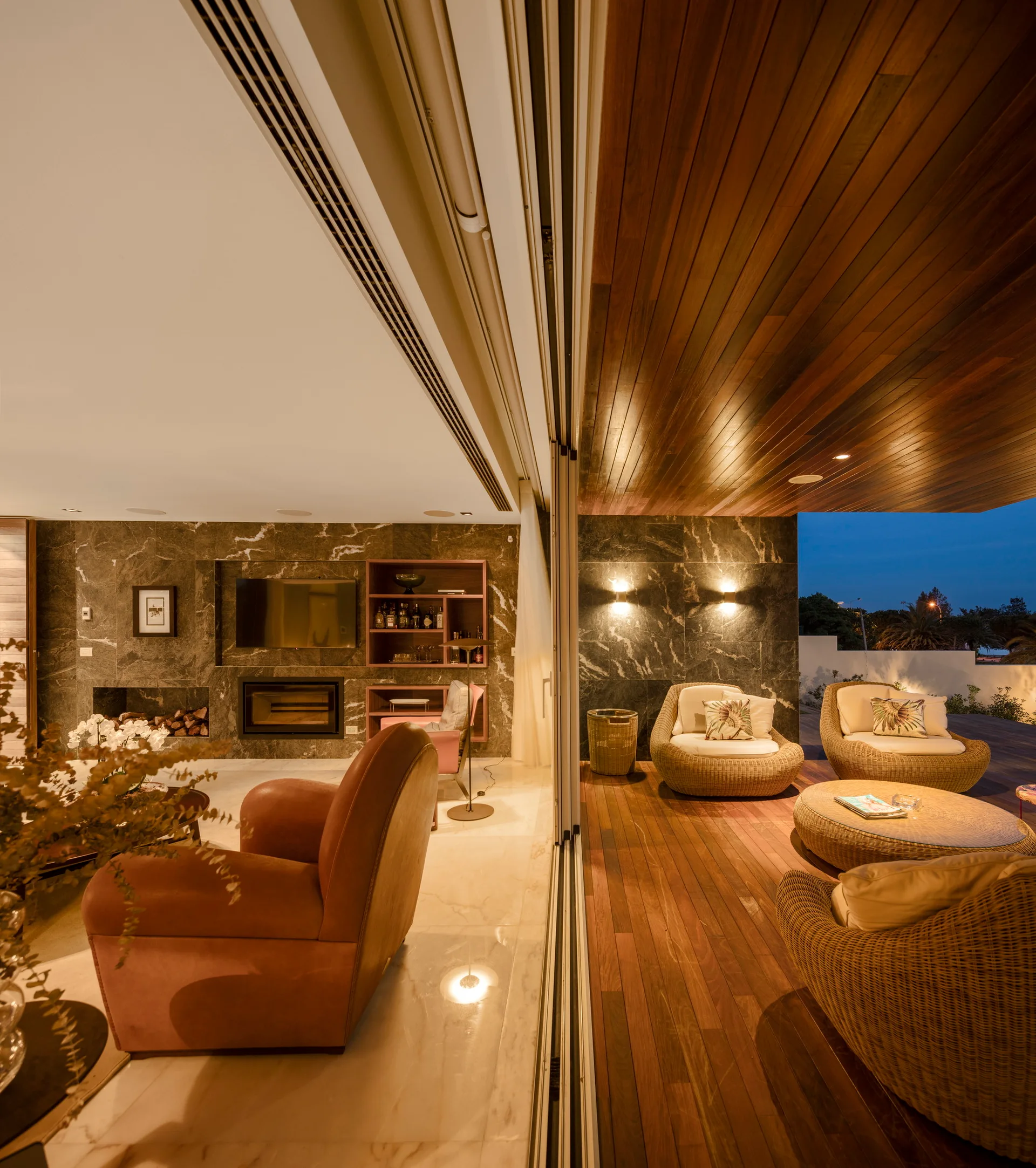A modern living room and a wooden patio deck are separated by open sliding glass doors, featuring dark veined stone walls and woven wicker furniture.