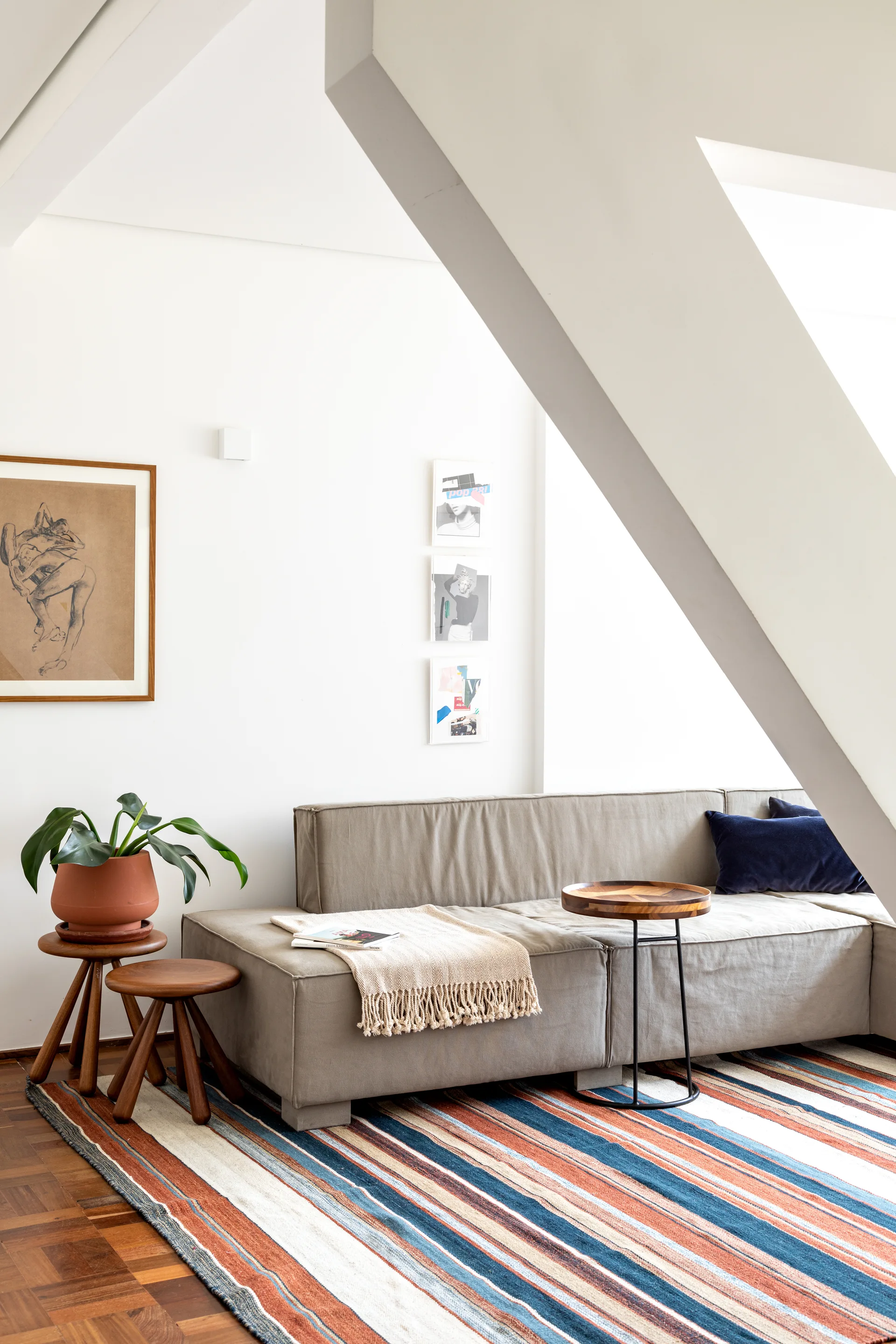 A low grey sectional sofa sits under a sloped white ceiling on a colorful striped rug in an attic living room with wood side tables.