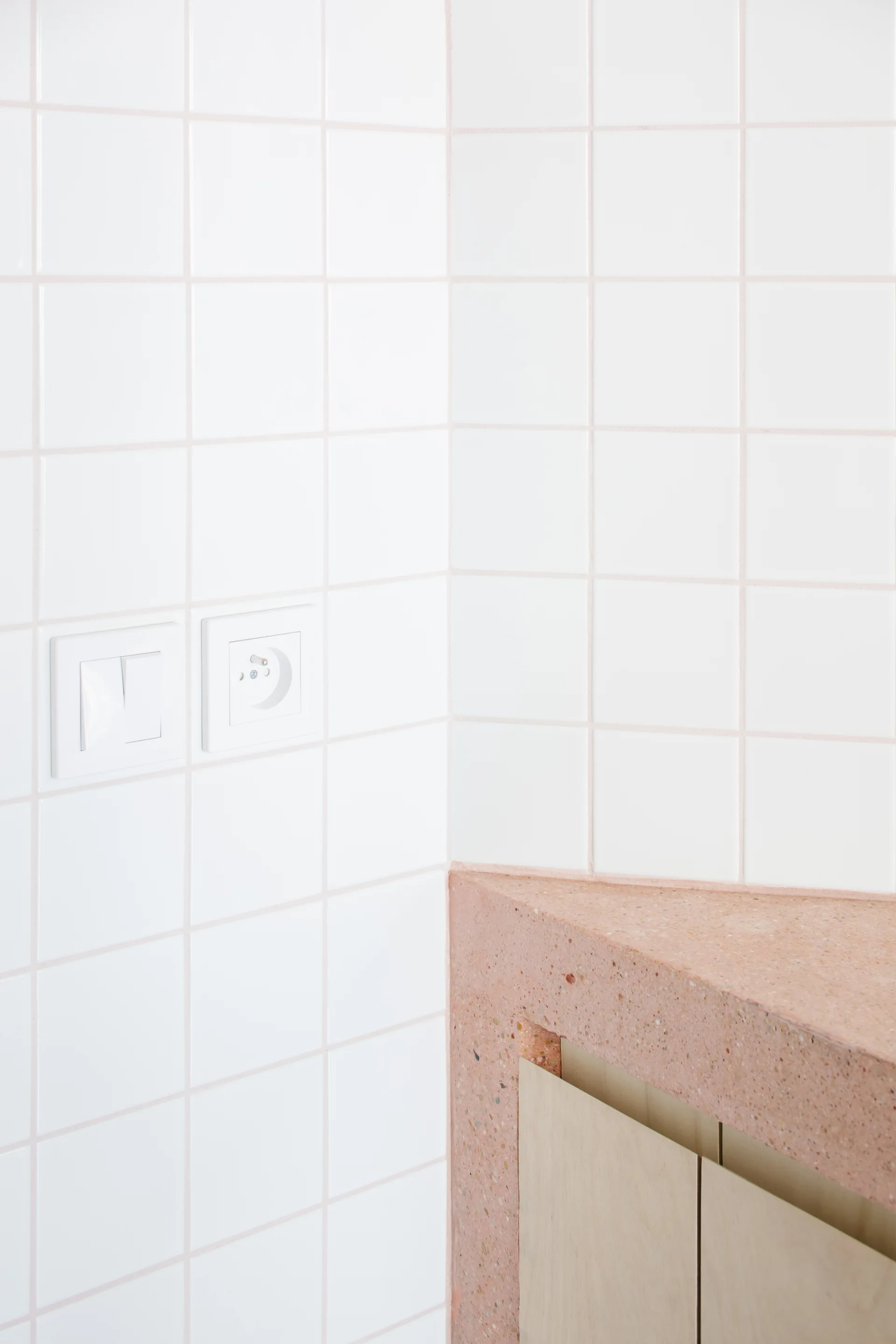 A corner wall of white square tiles with a light switch and outlet sits above a pink terrazzo countertop and light wood cabinets.