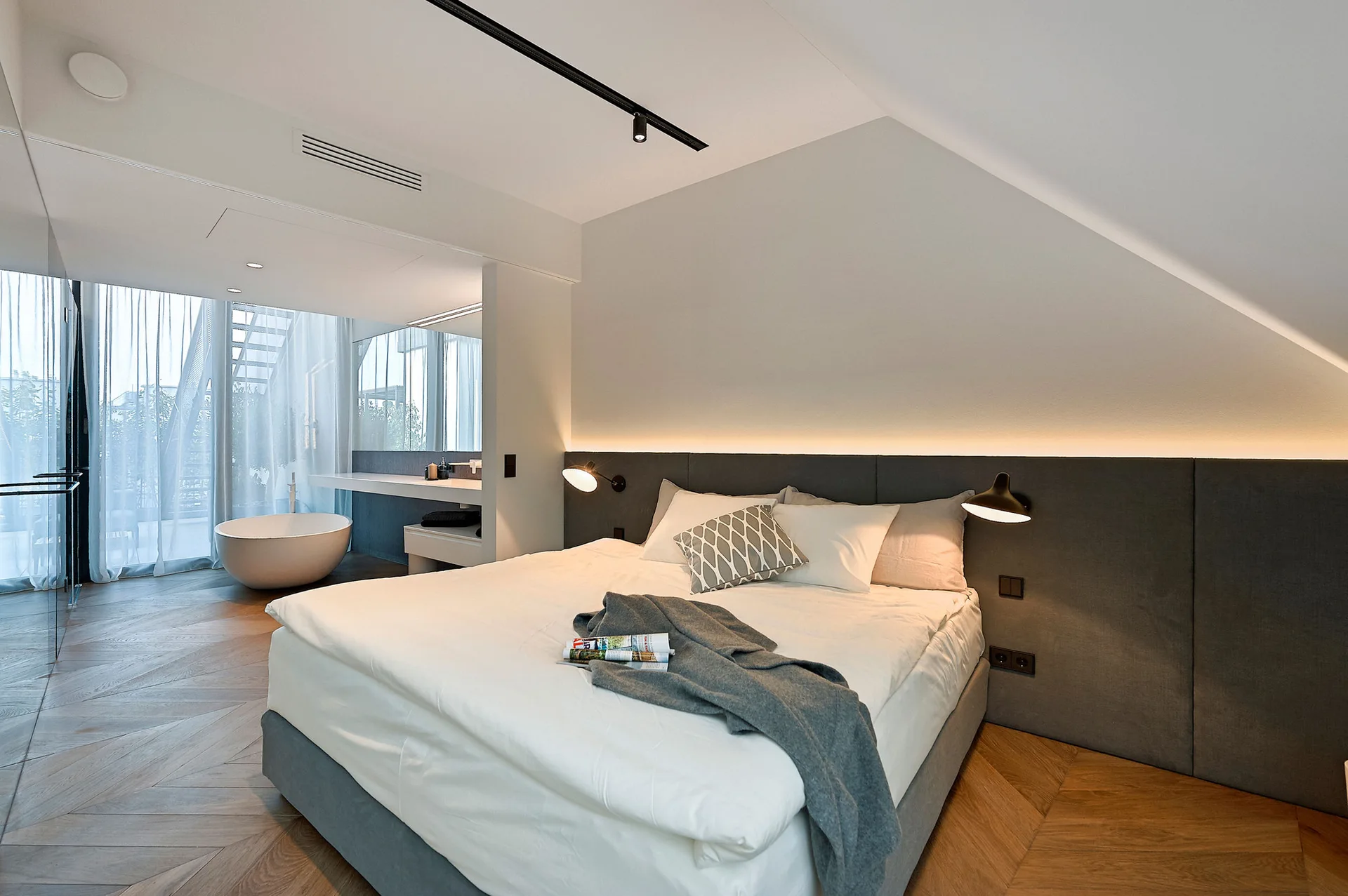 A modern bedroom under a sloped ceiling features a grey upholstered headboard, chevron wood flooring, and an open-plan bathroom with a freestanding tub.