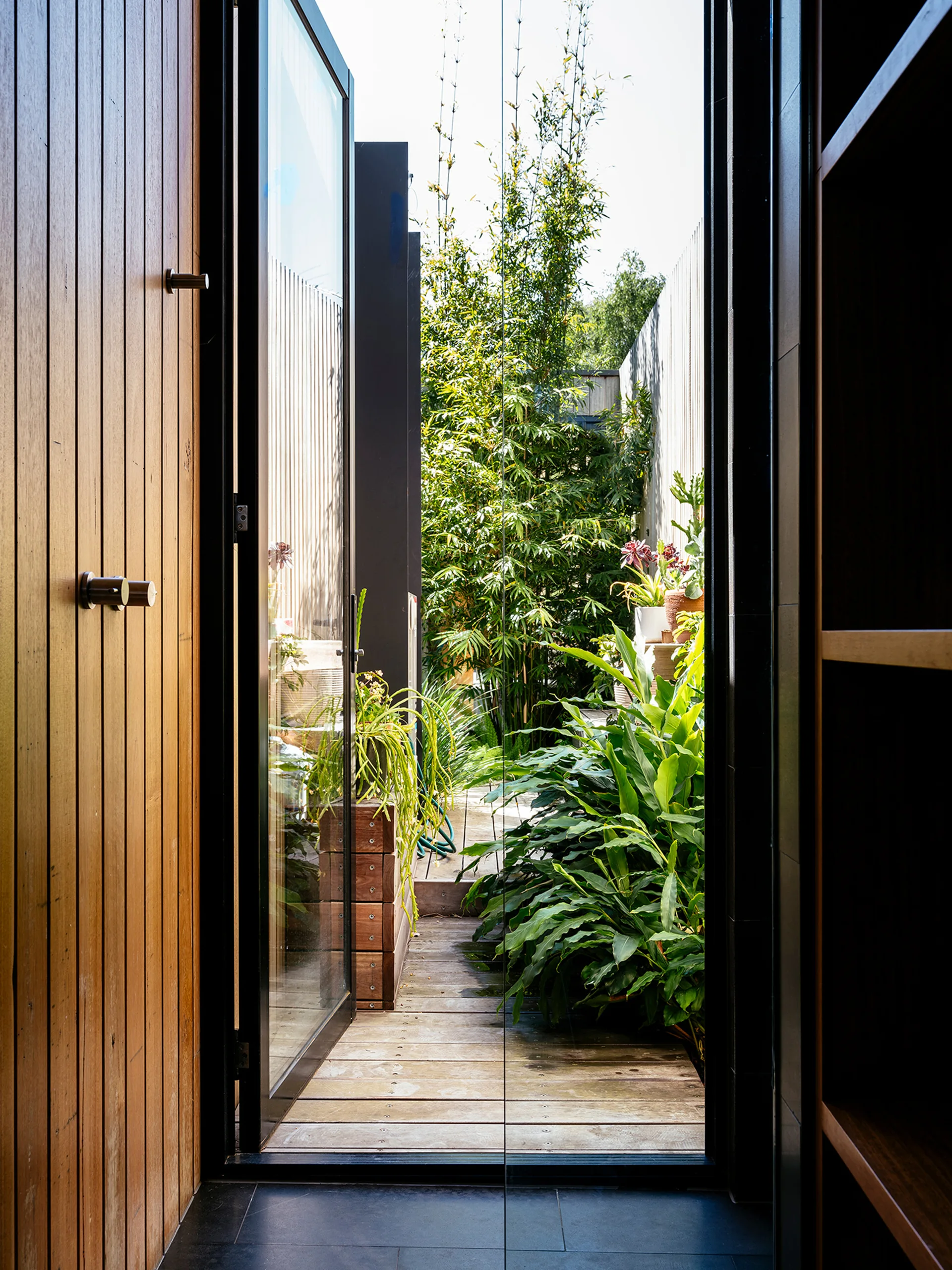 A tall black-framed glass door opens from a wood-paneled interior onto a small outdoor courtyard with weathered wood decking and leafy green plants.