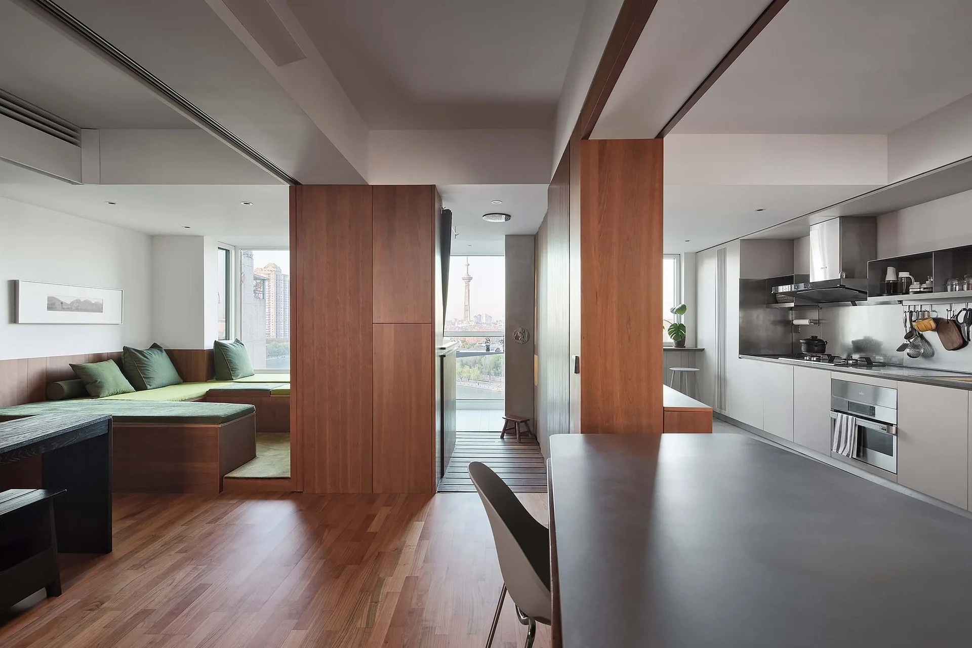 An open-plan modern apartment featuring a raised wooden platform lounge with green cushions, a central wood-paneled partition, and a sleek kitchen.