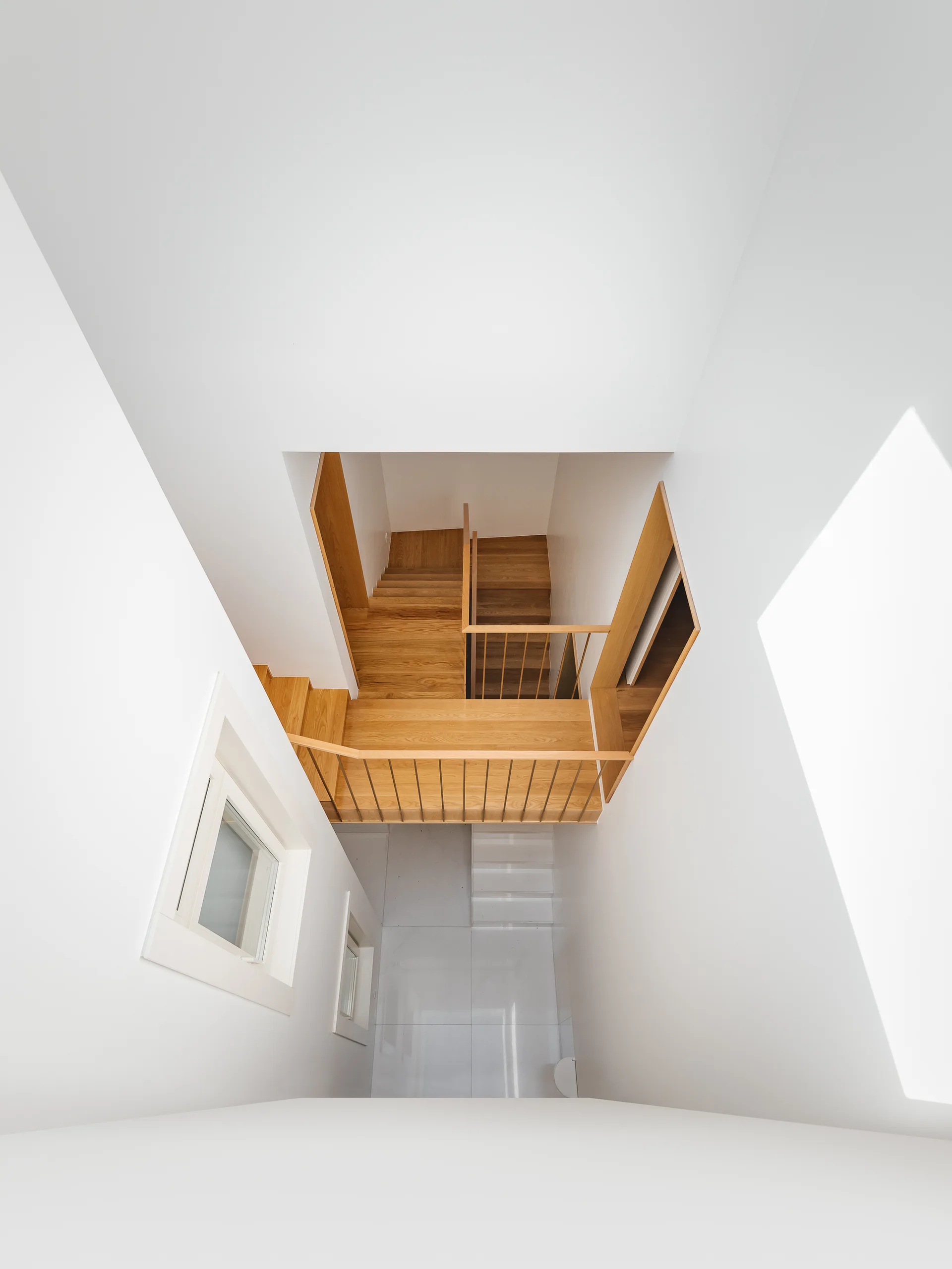 A high-angle view looking down a minimalist white stairwell featuring warm wood stairs and landings with thin metal railings.