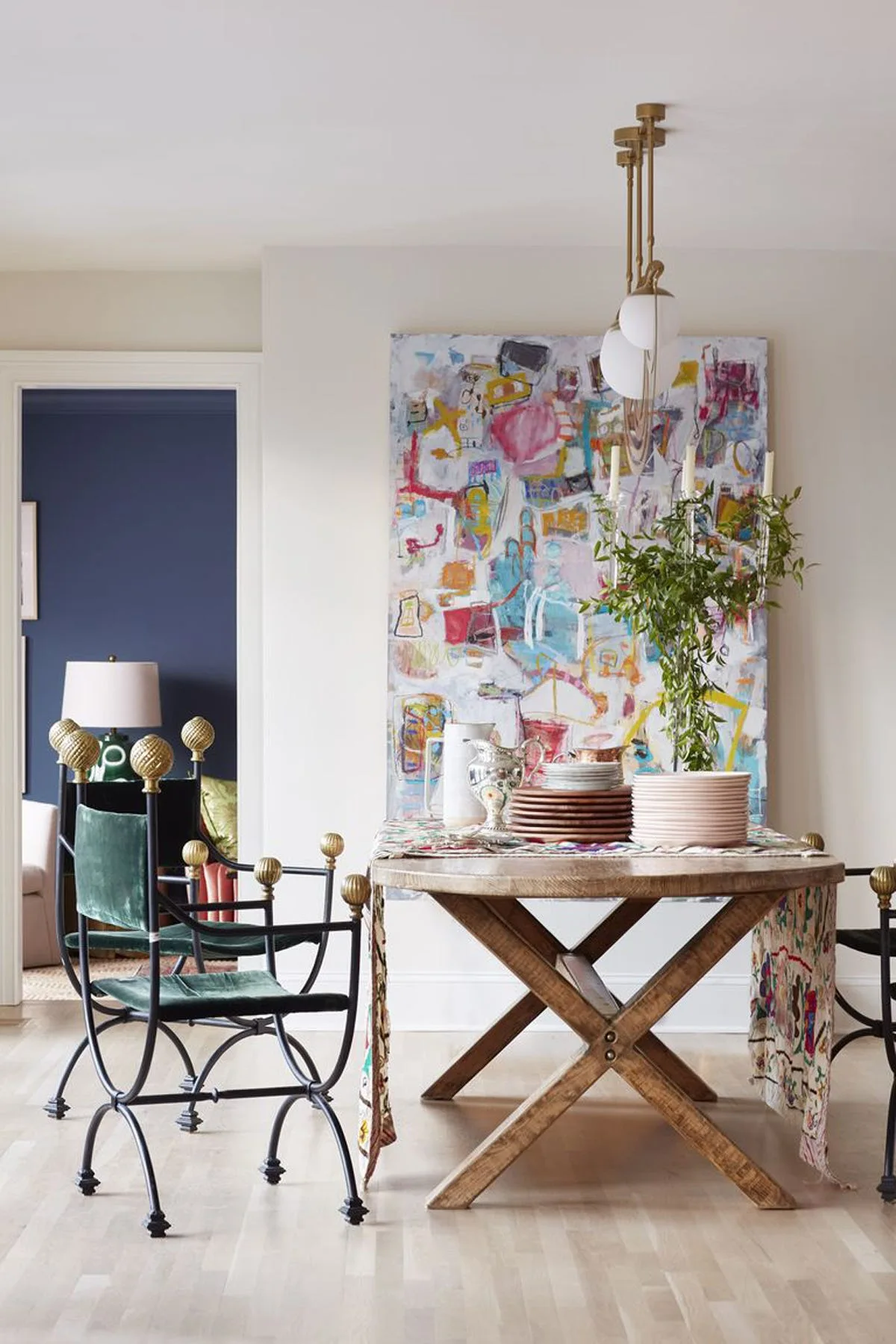 An eclectic dining room featuring a wooden trestle table, green velvet chairs with gold finials, and a large colorful abstract painting.