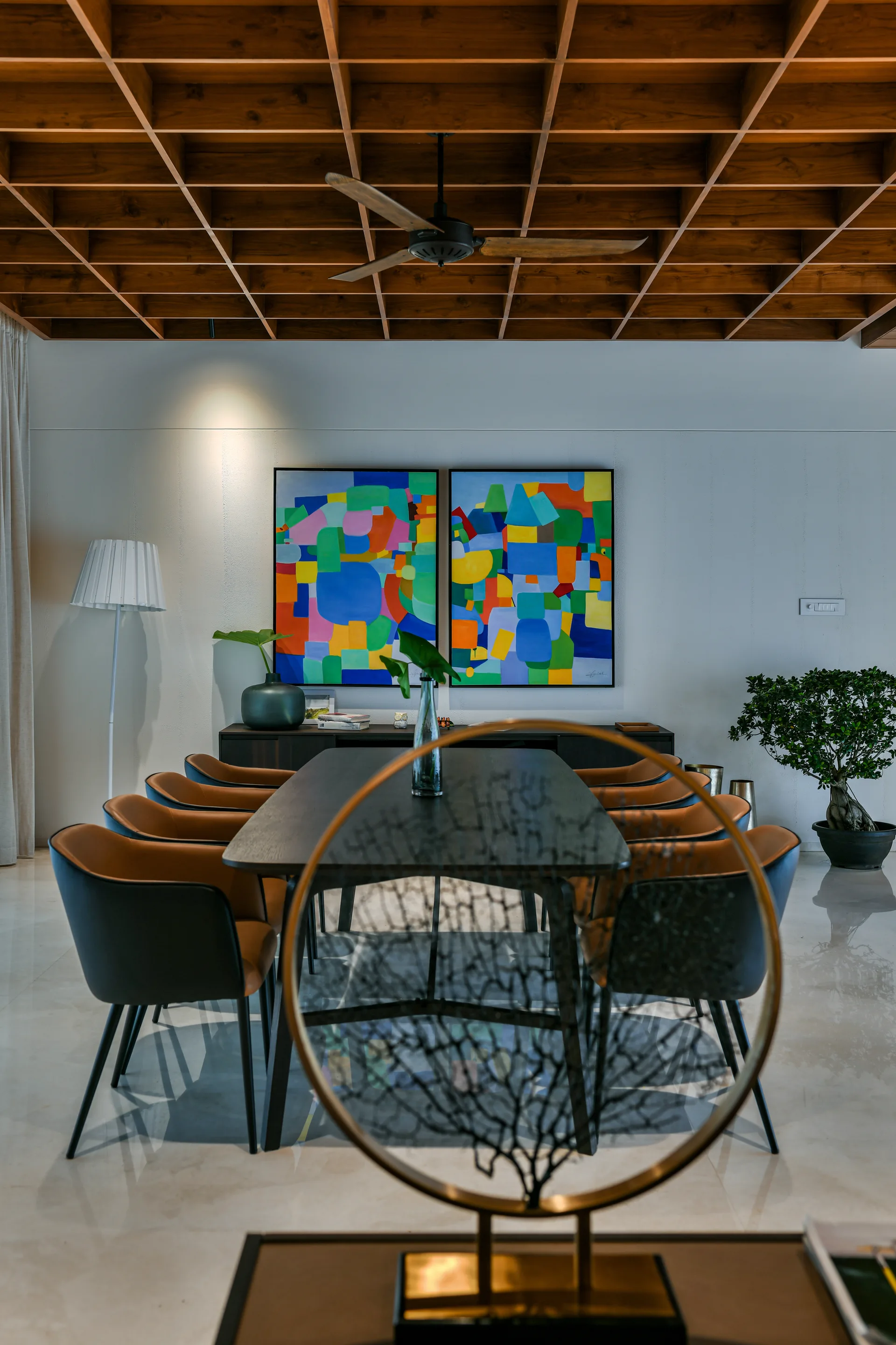 A modern dining room with a dark table, leather-lined chairs, a wooden grid coffered ceiling, and two colorful abstract paintings on the back wall.