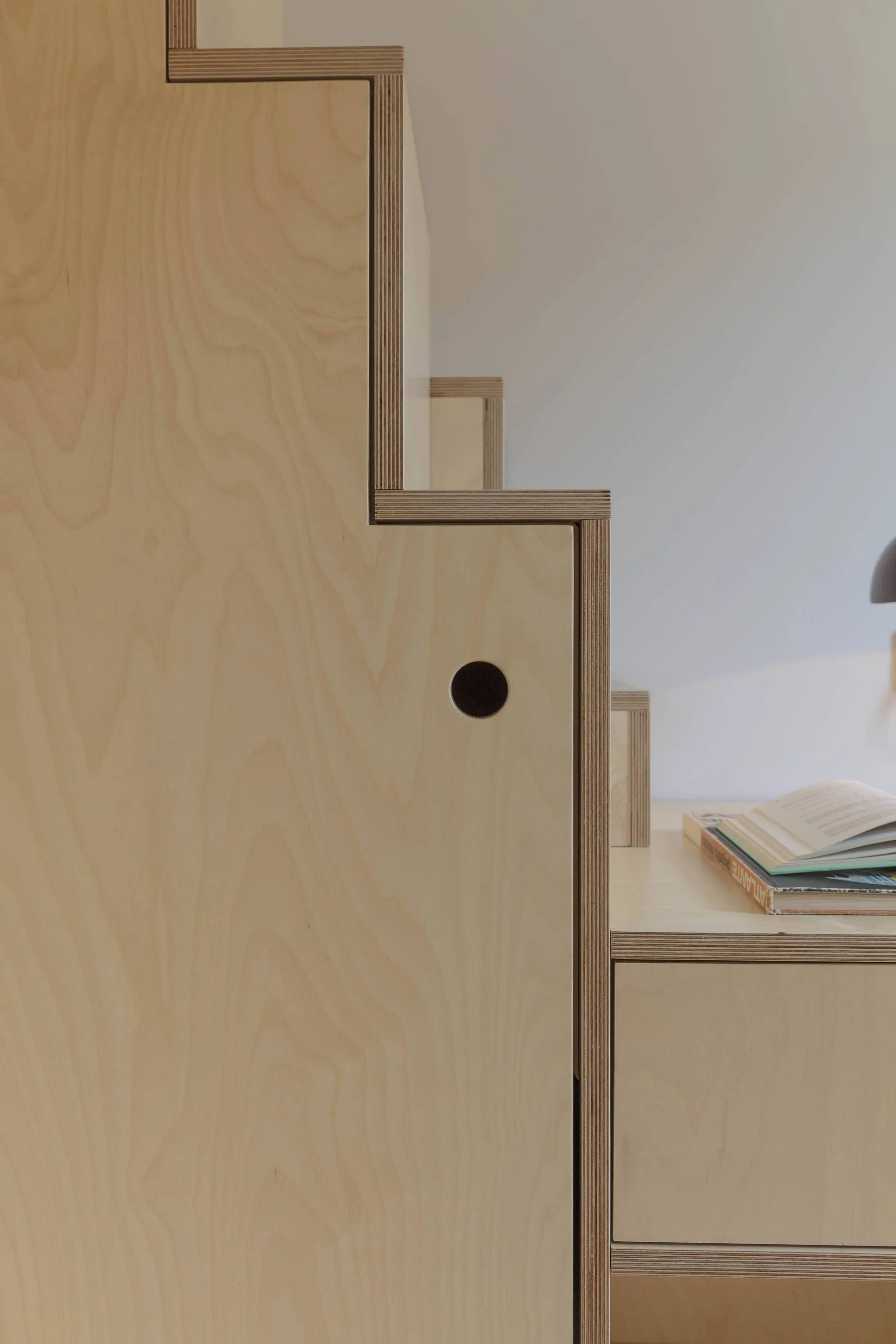 A close-up of light-toned plywood stairs featuring exposed layered edges, integrated storage compartments, and a circular finger pull.