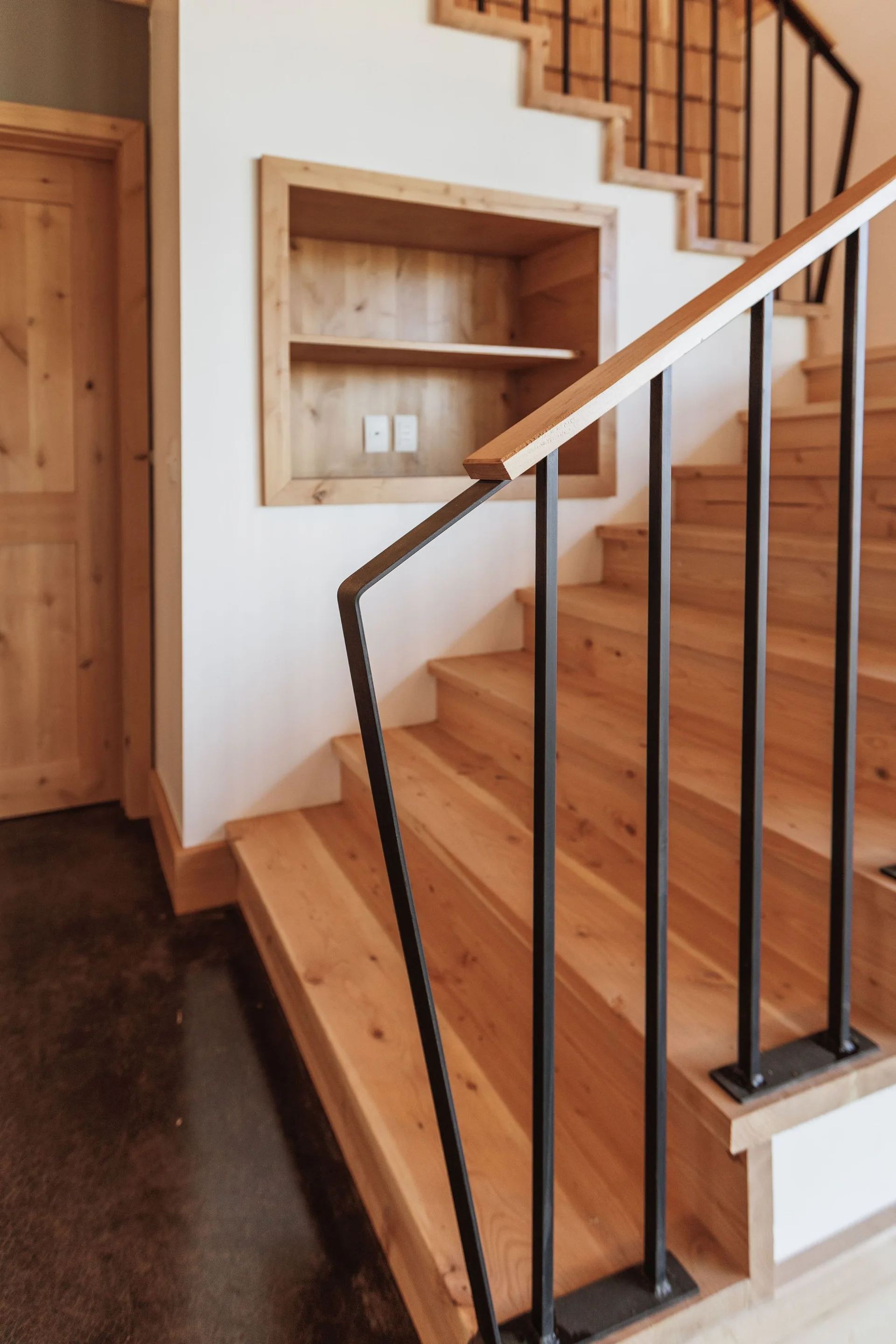 A wooden staircase with a black metal handrail stands next to a white wall containing a recessed wooden shelving unit.
