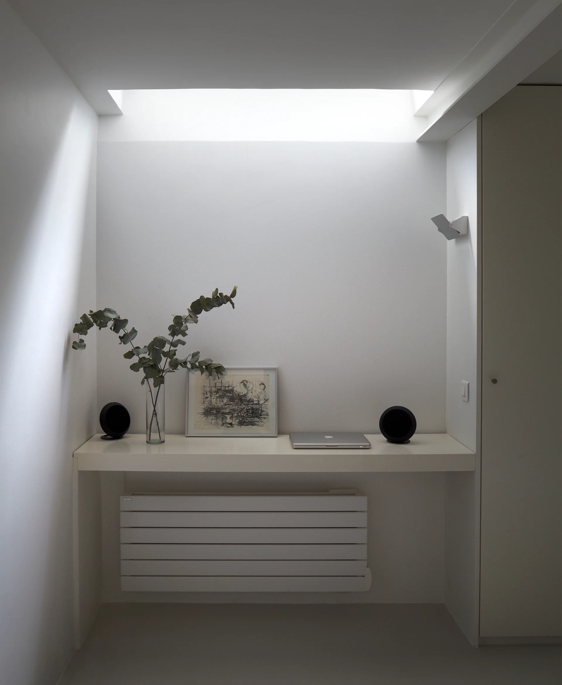 A minimalist white built-in desk under a bright skylight, featuring a laptop, speakers, and a plant in a small alcove.