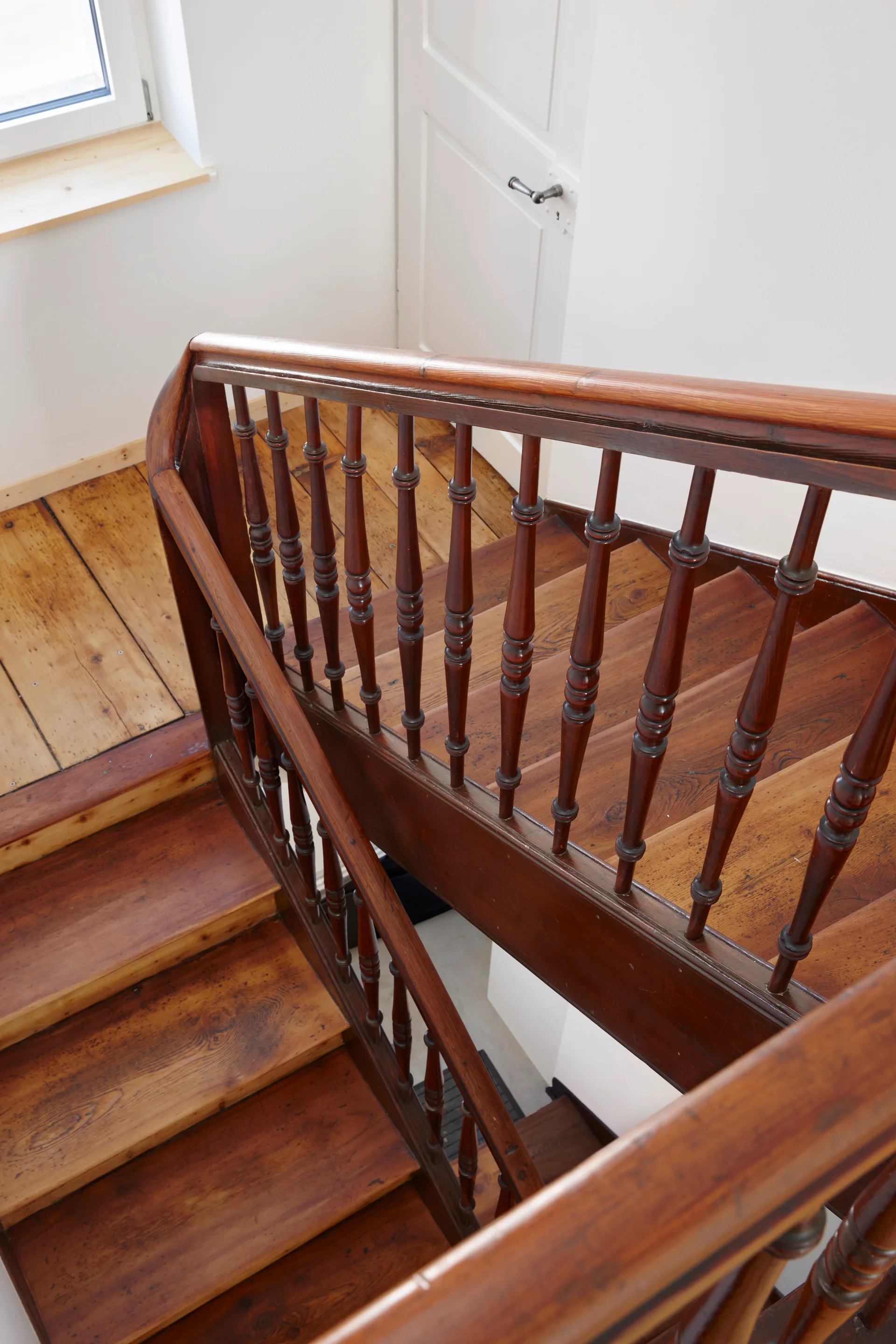 A high-angle view of a wooden staircase with stained wood steps, a curved handrail, and classic turned balusters against white walls.