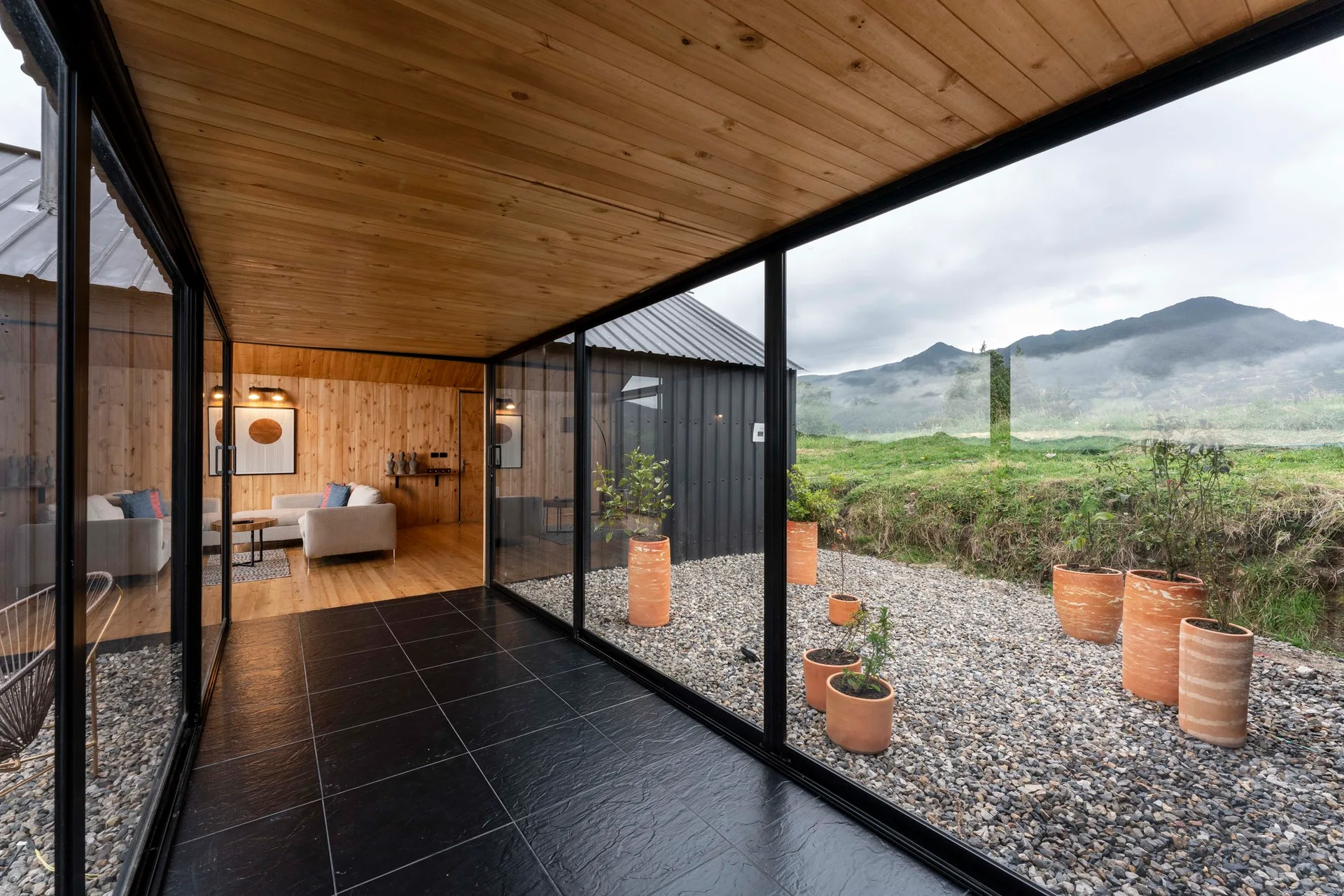 A modern glass-walled hallway with black tiled floors overlooks a gravel courtyard with terracotta planters, showing a wood-paneled living room inside.
