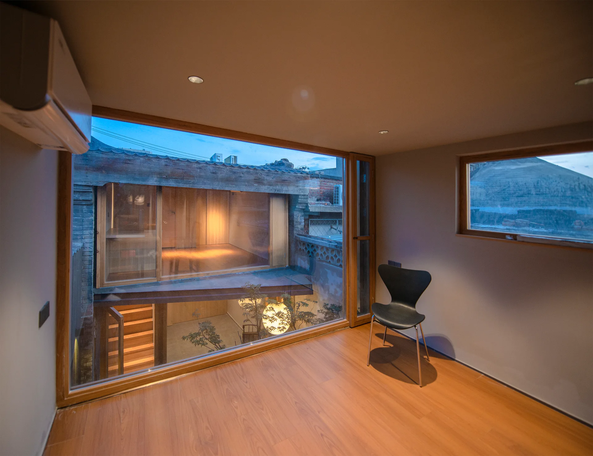 A minimalist room features wood floors, a black chair, and a large window wall overlooking a multi-level courtyard.