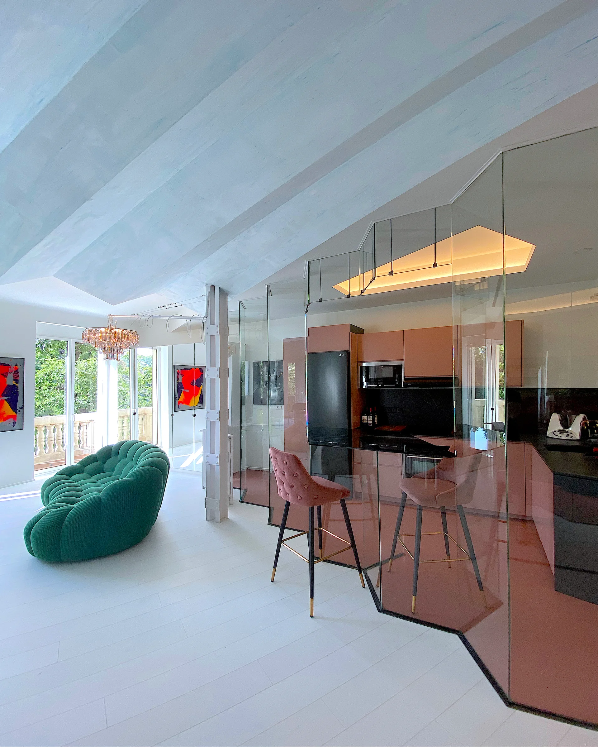 A contemporary open-plan interior with a curved green tufted sofa and a pink kitchen enclosed behind an angled glass partition wall.