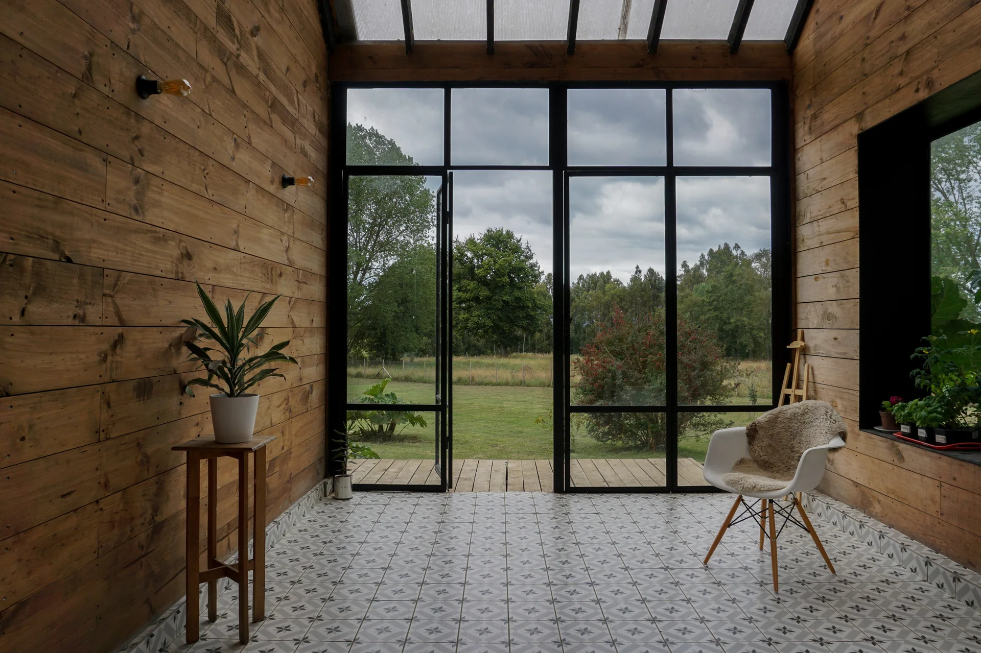 A sunroom featuring horizontal wood plank walls, geometric patterned floor tiles, and a large black metal framed glass door opening to a garden.