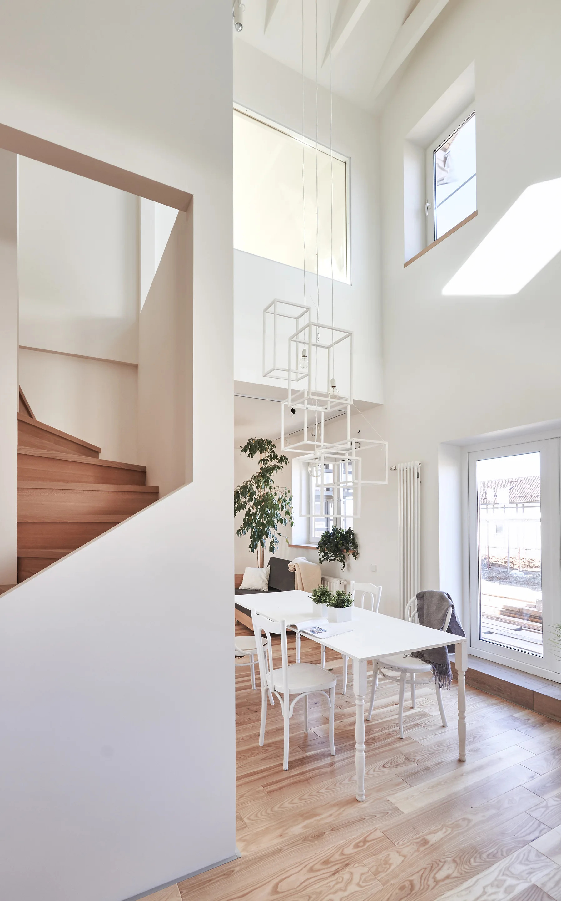 A bright double-height dining room features a white wooden table and chairs, a geometric frame pendant light, and a natural wood staircase.