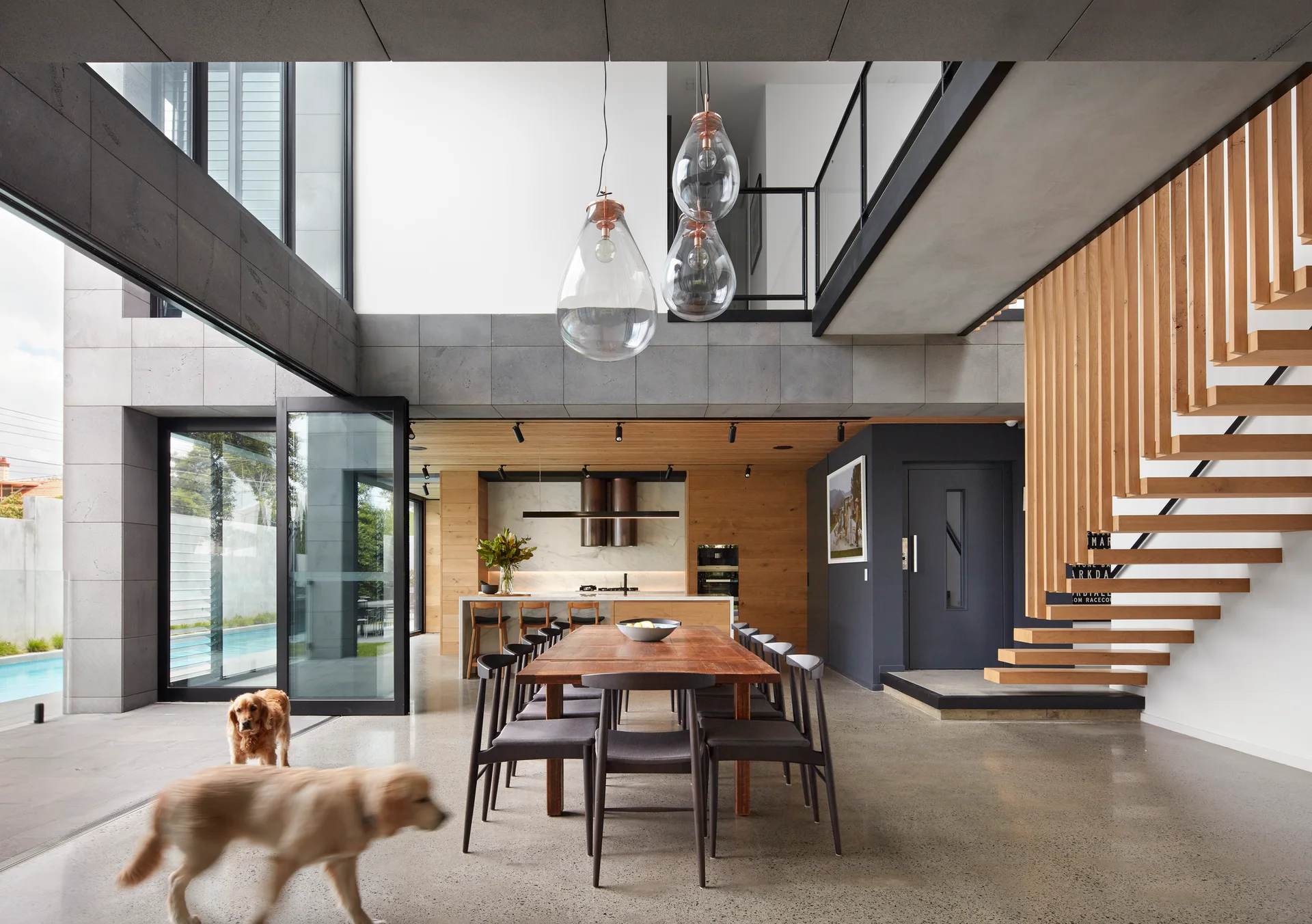 A modern open-plan dining room features a long wooden table, a floating timber staircase, and large glass pendant lights next to glass sliding doors.
