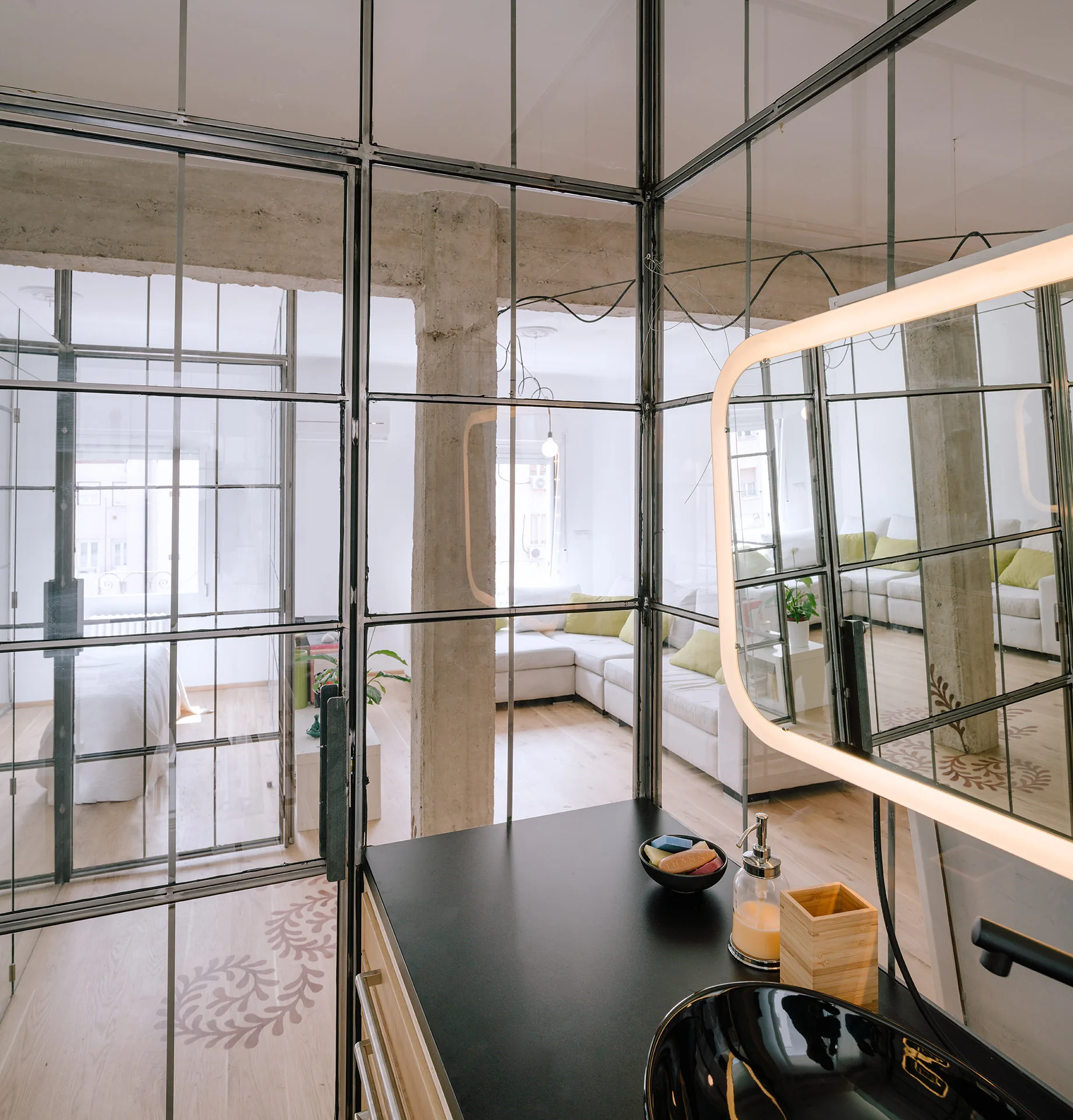 A modern bathroom vanity with a black vessel sink and illuminated mirror is enclosed by industrial black metal-framed glass partition walls.
