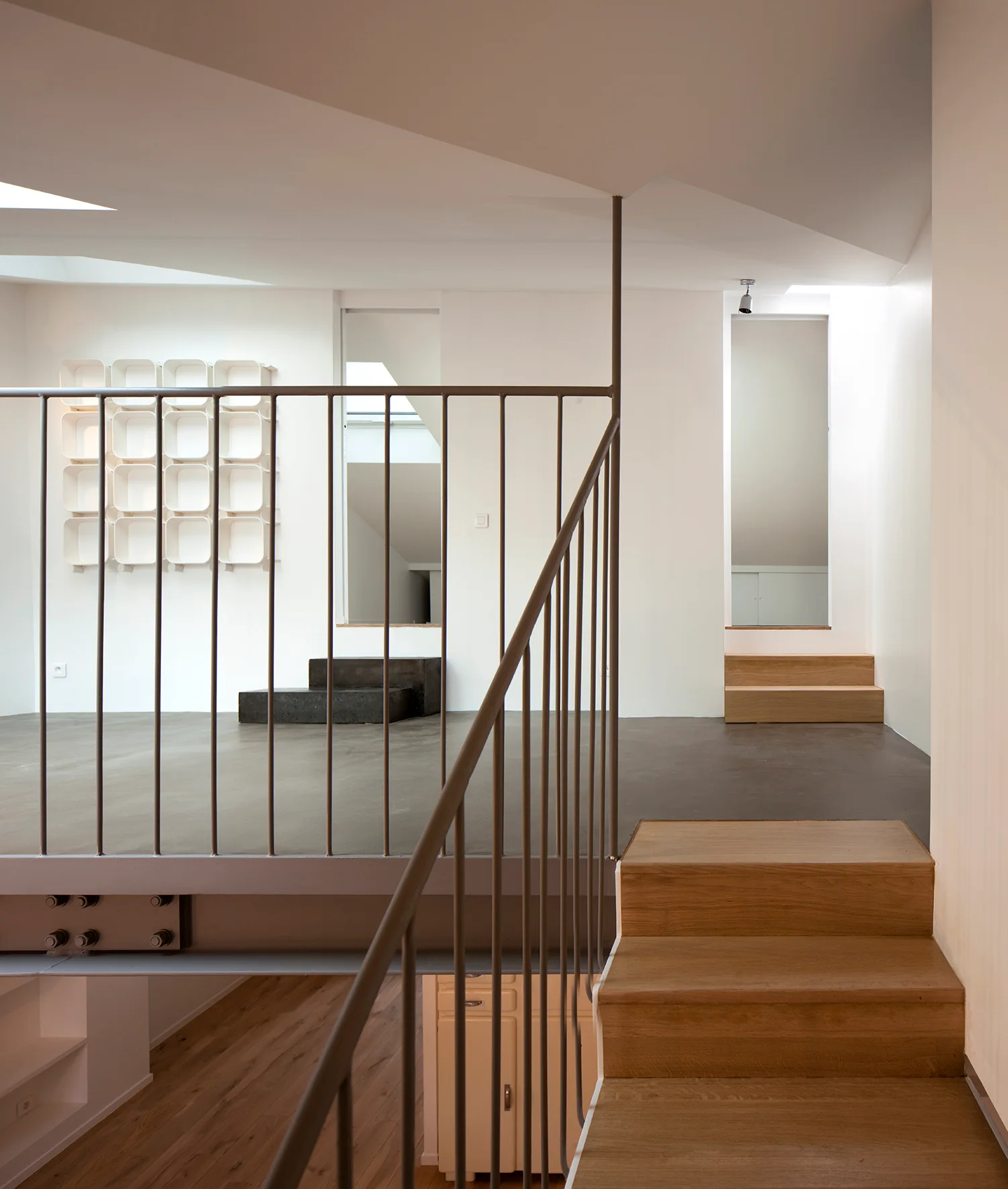 A split-level loft space featuring a wooden staircase, a slender grey metal railing, and a minimalist grid wall shelf against white walls.