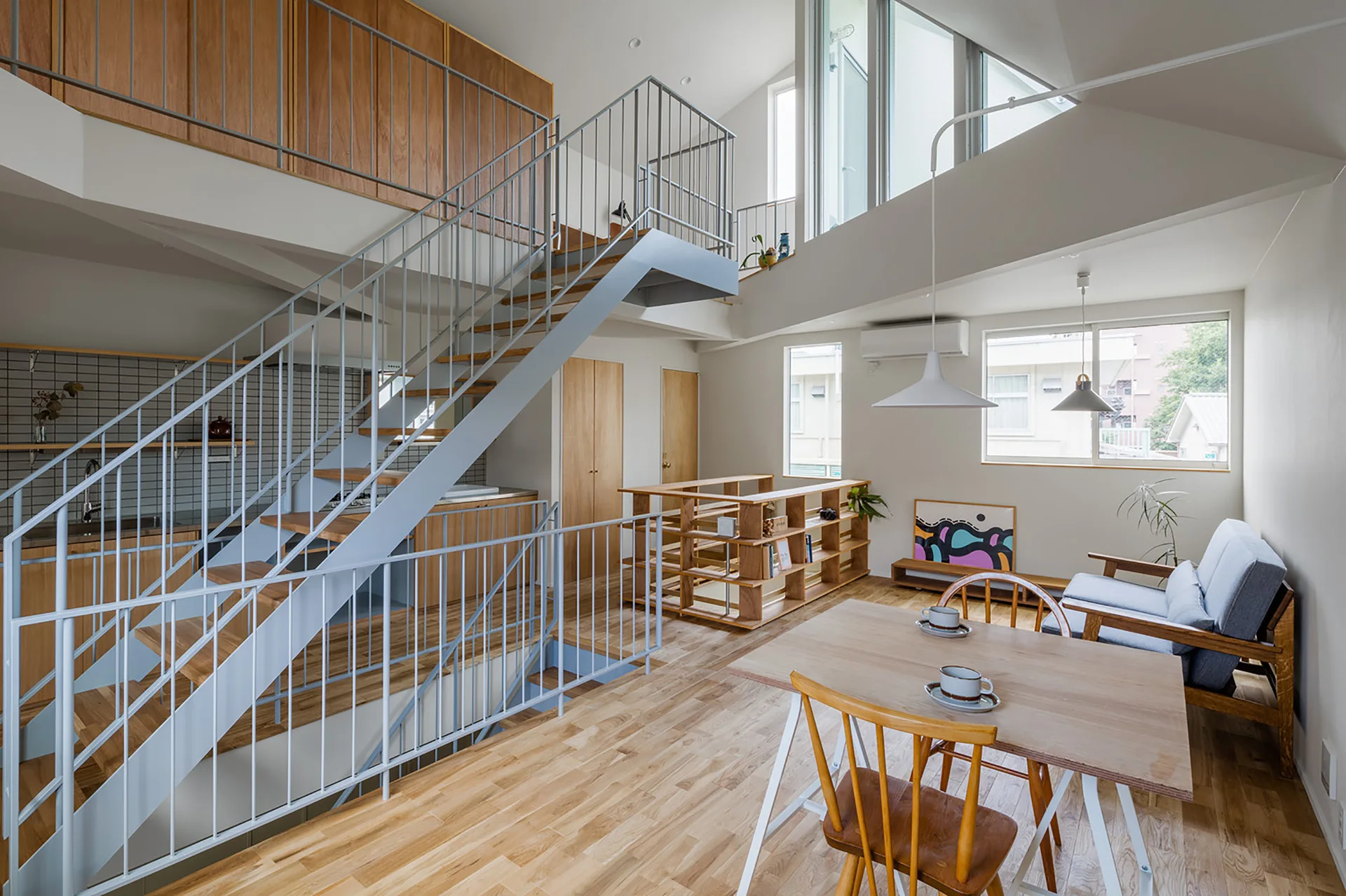 An open-plan living and dining area featuring a light wood dining set, a gray sofa, and a central gray metal staircase leading to a mezzanine.