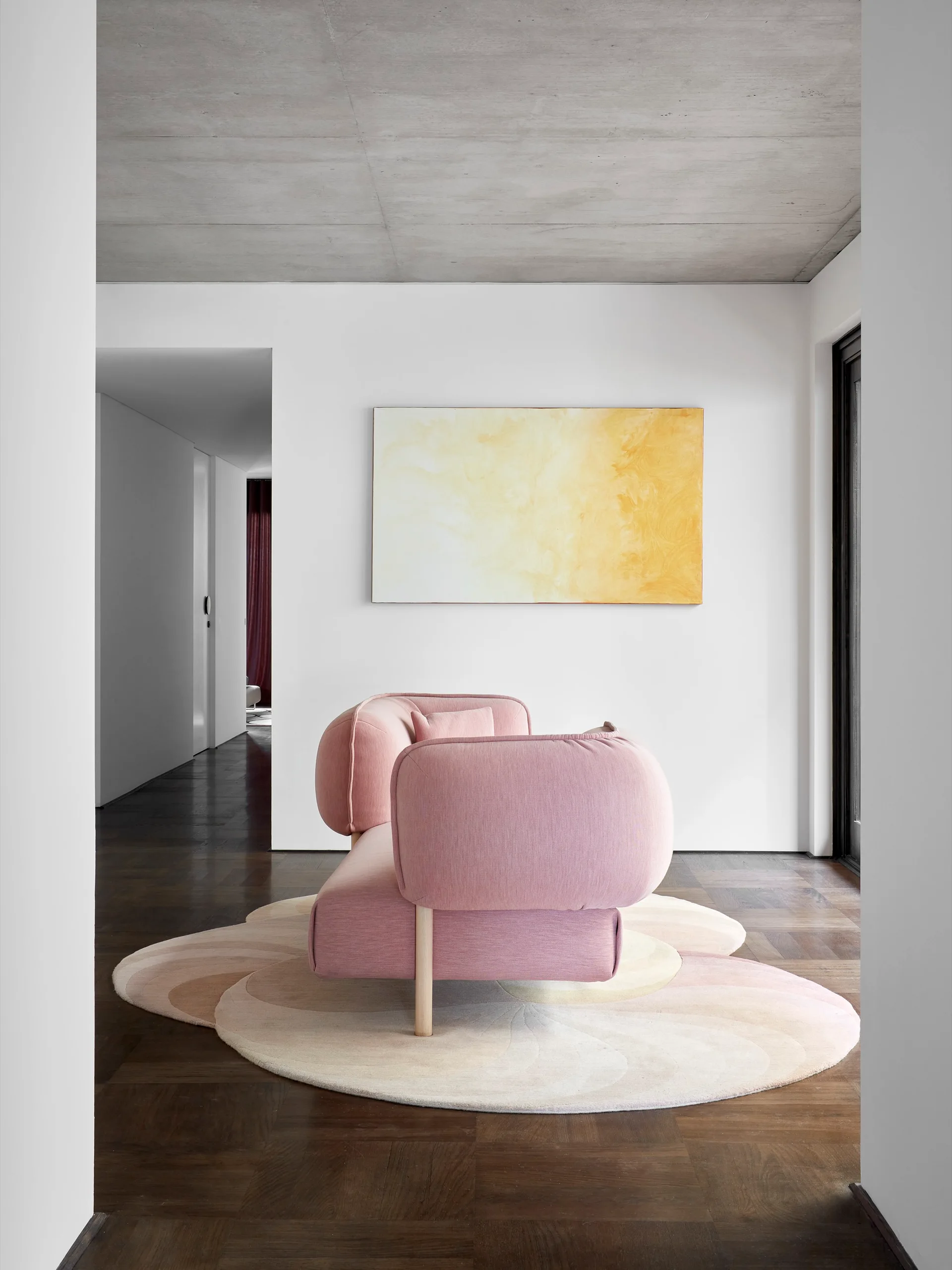 A modern pink sofa sits on a curved, layered pastel rug on dark wood parquet flooring beneath an exposed concrete ceiling.
