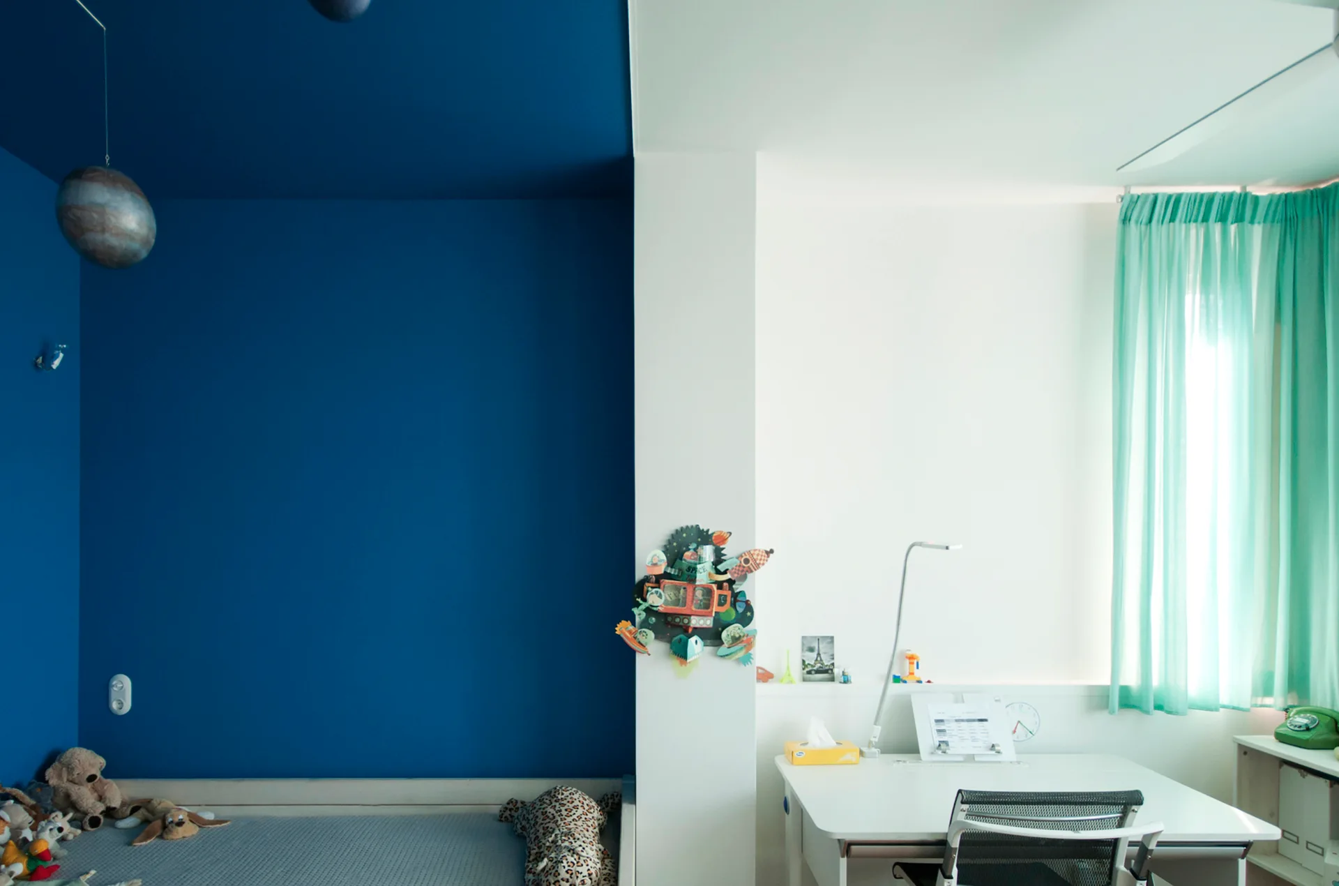 A children's bedroom is split into a dark blue sleeping zone with a bed and a bright white study zone with a desk and teal curtains.