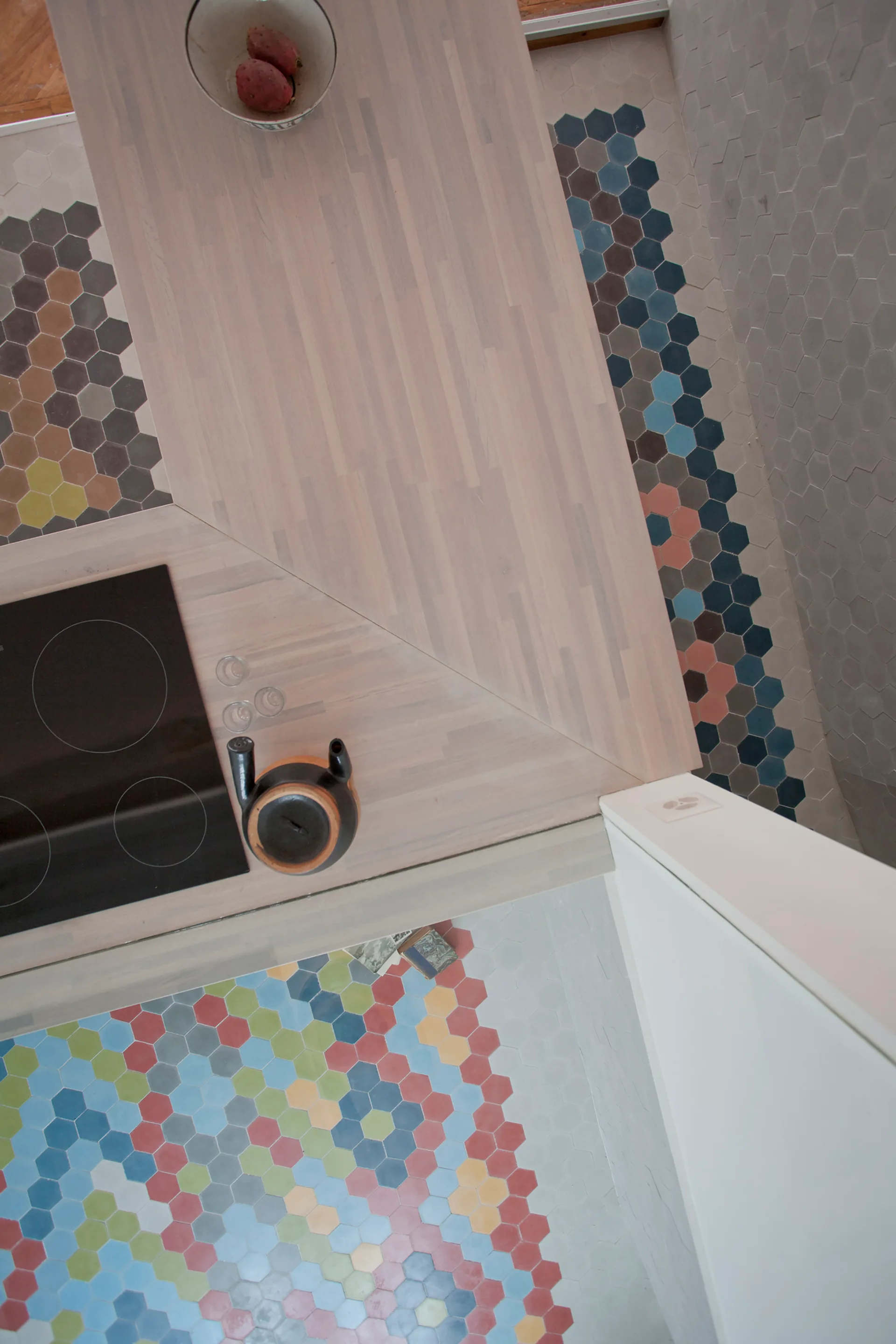 An overhead view of a light wood kitchen countertop with an induction stove and colorful hexagonal floor tiles below.