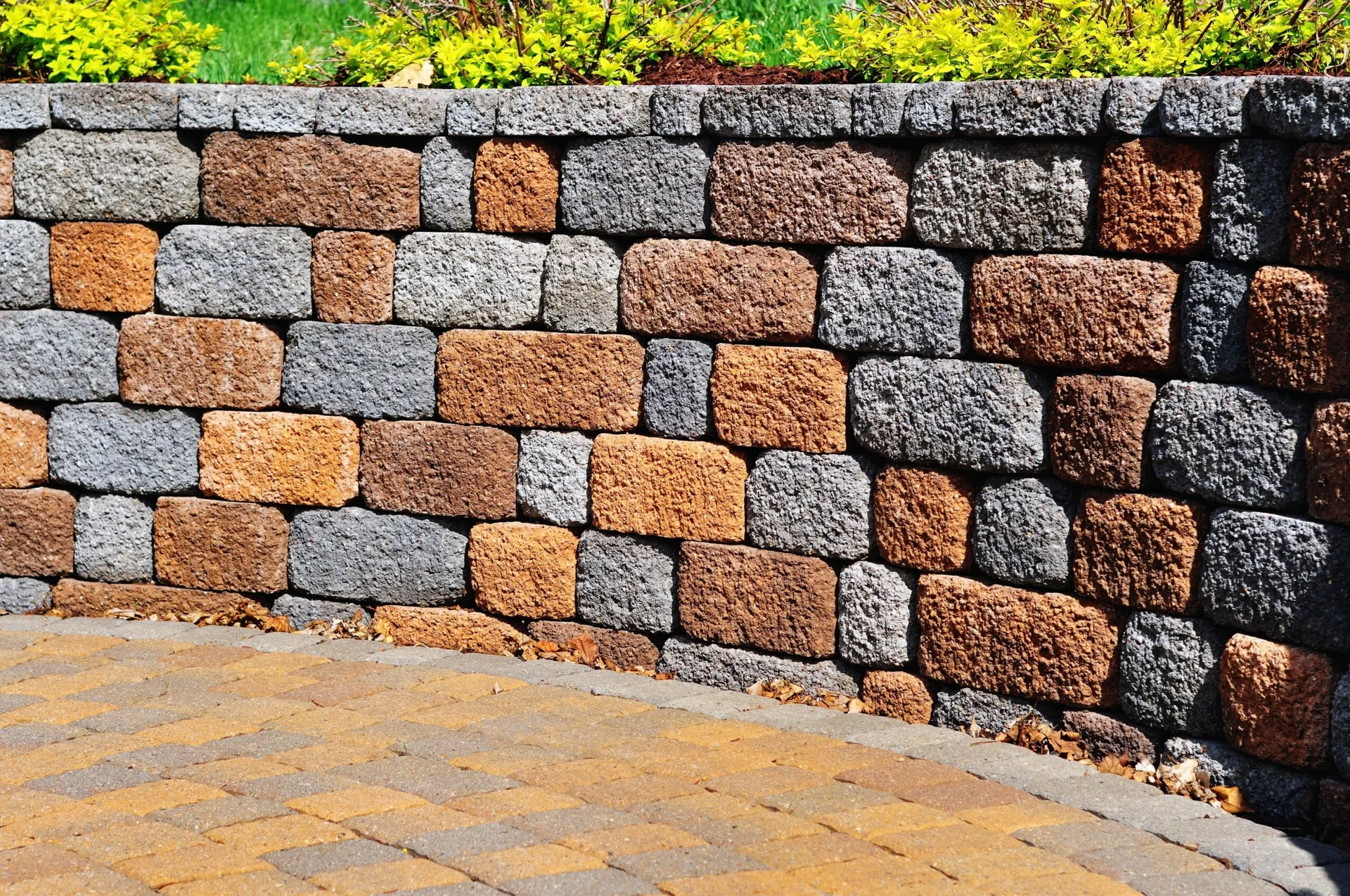 A curved retaining wall made of textured grey and rust stone blocks borders a paved outdoor patio.