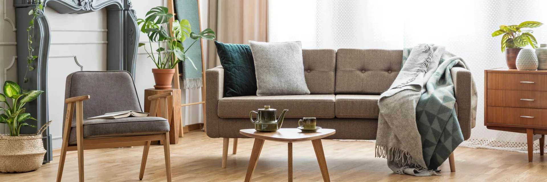A bright living room features a mid-century modern sofa, a wooden coffee table, a gray armchair, and potted houseplants against a white wall.