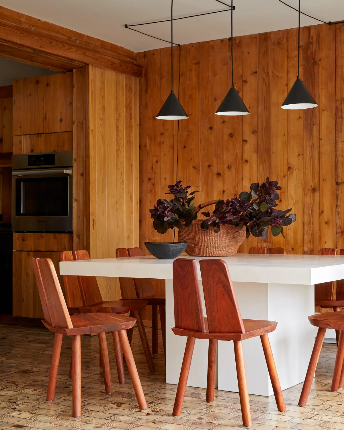 A white square dining table with sculptural wooden chairs is set against vertical wood-paneled walls beneath three black pendant lights.