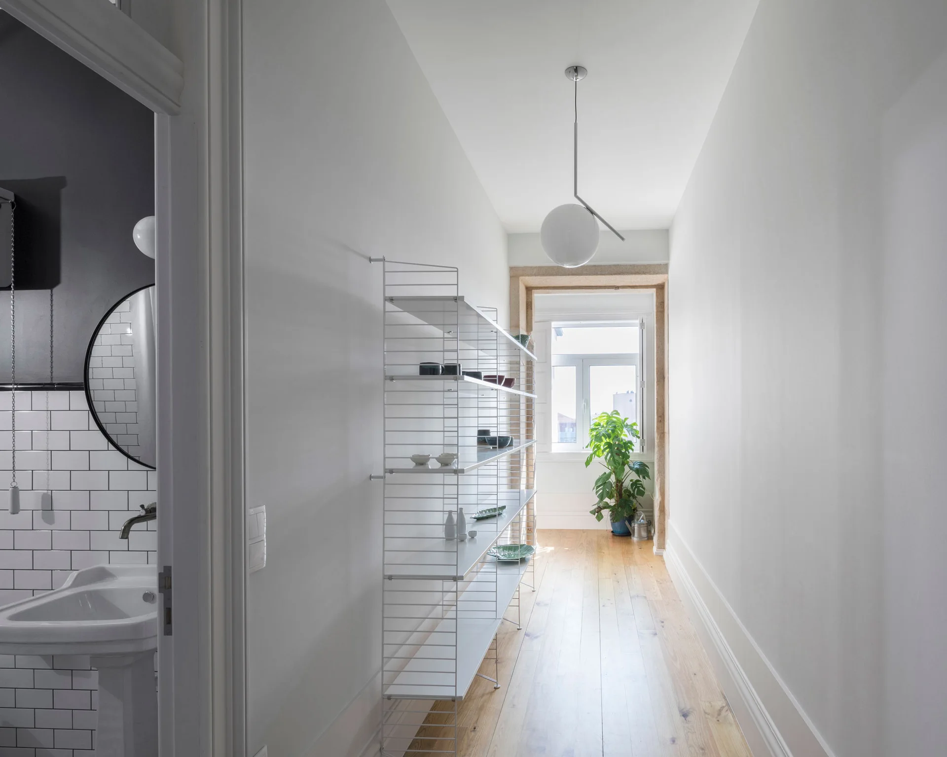 A bright hallway features light wood floors, a tall white wire shelving unit, a hanging globe pendant light, and an open doorway to a bathroom.