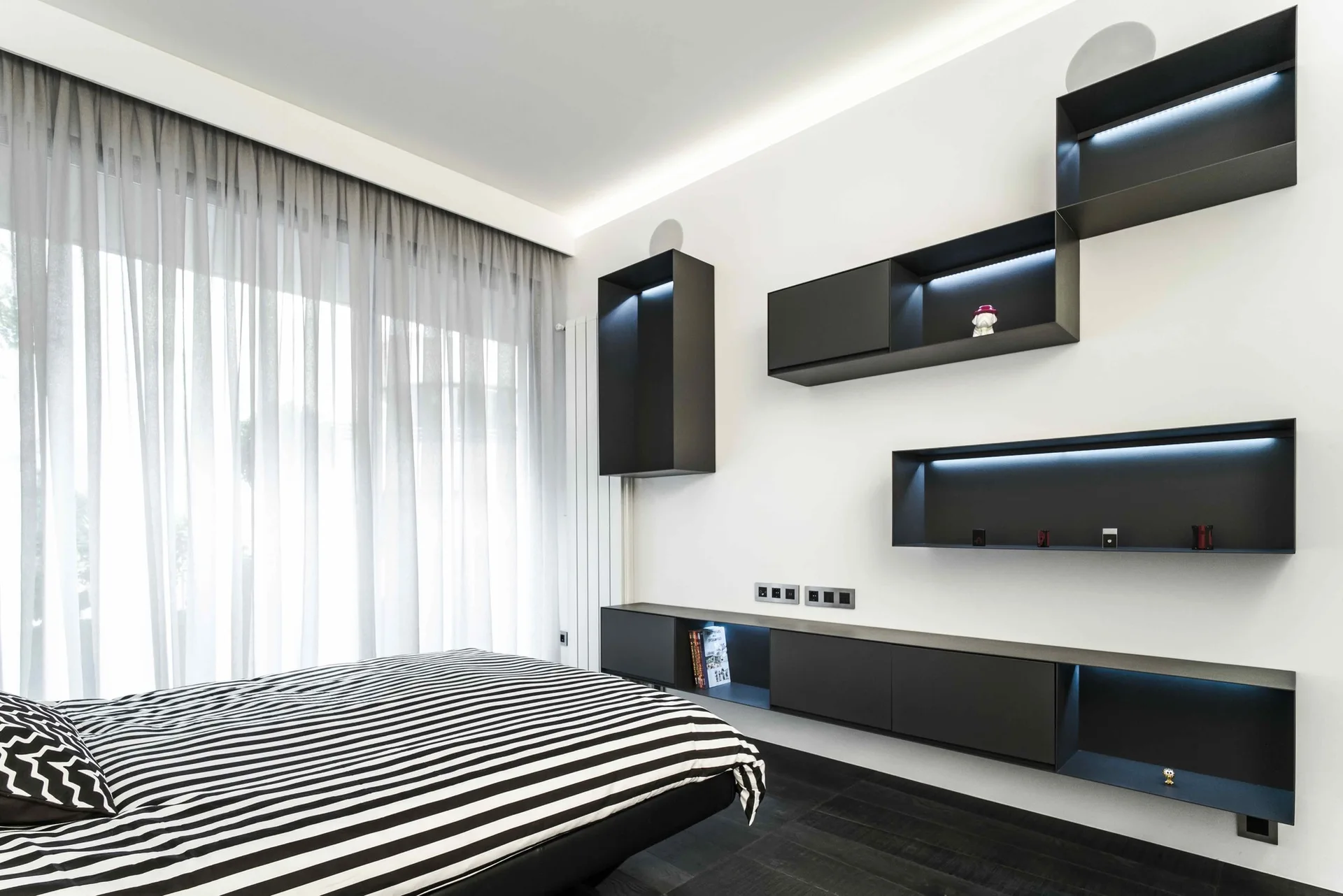 A modern bedroom features a bed with black and white striped bedding, sheer curtains, and staggered black geometric shelves with built-in LED lighting.