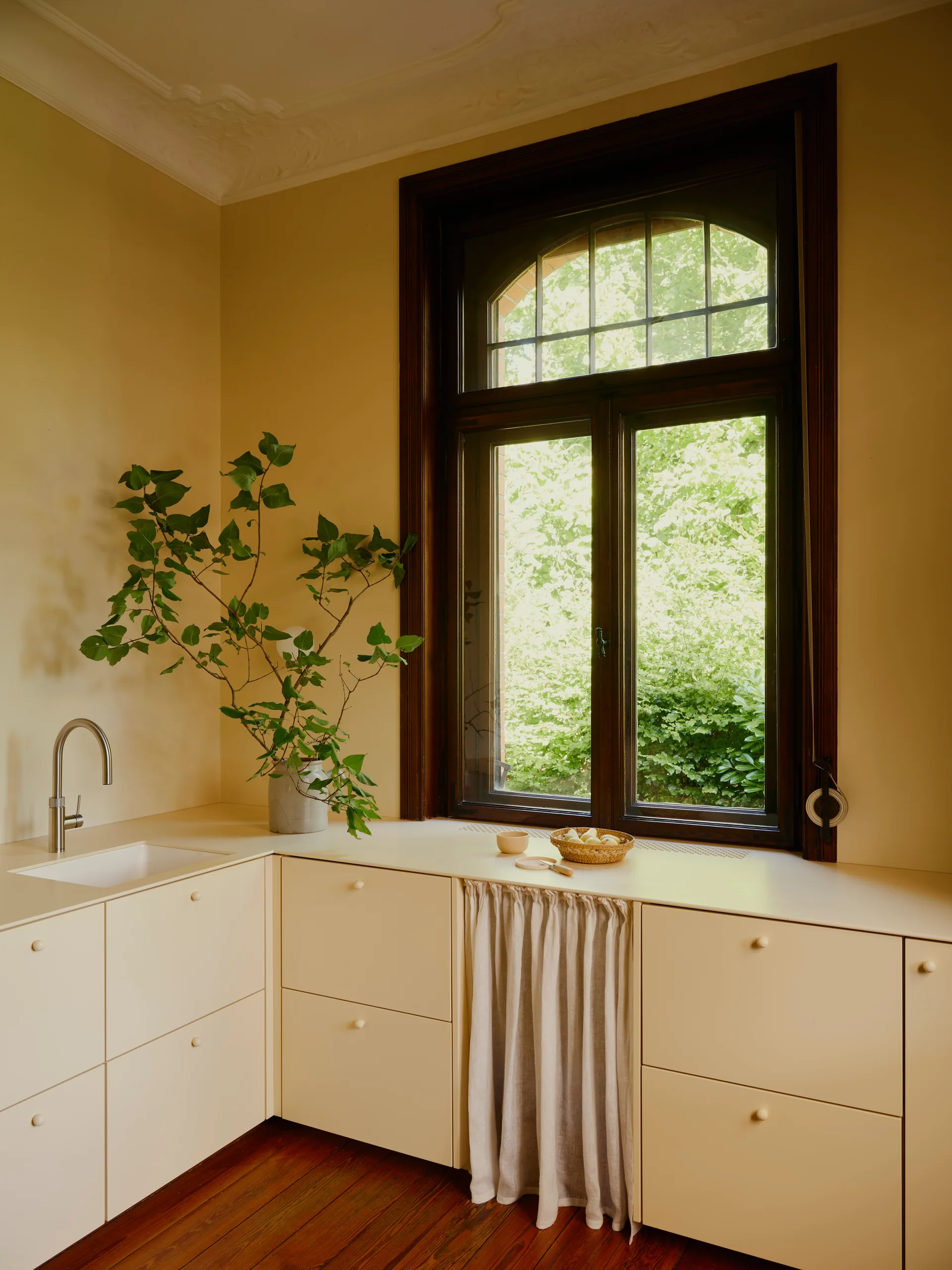 Cream-colored kitchen cabinets line a corner below a large, dark wood arched window looking out onto green foliage.