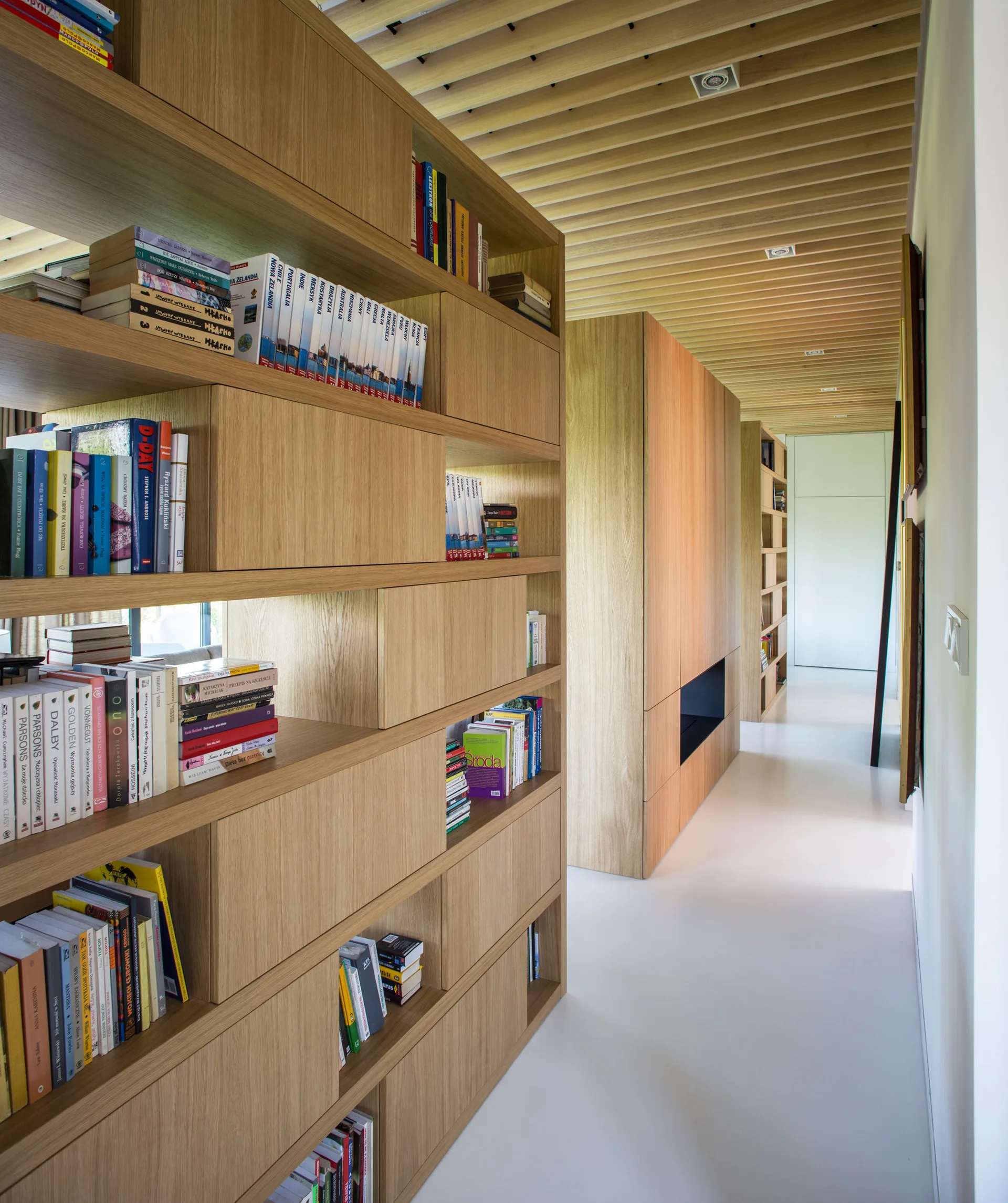 A modern white hallway featuring a large light wood built-in bookcase and a matching slatted timber ceiling with recessed lighting.