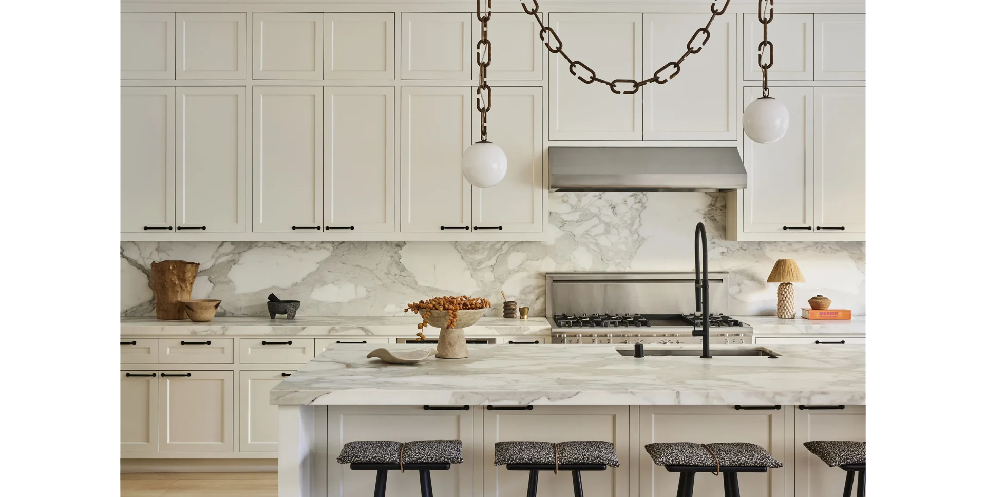 Cream kitchen cabinetry frames a veined marble backsplash and island, illuminated by globe pendant lights hanging from rustic metal chains.