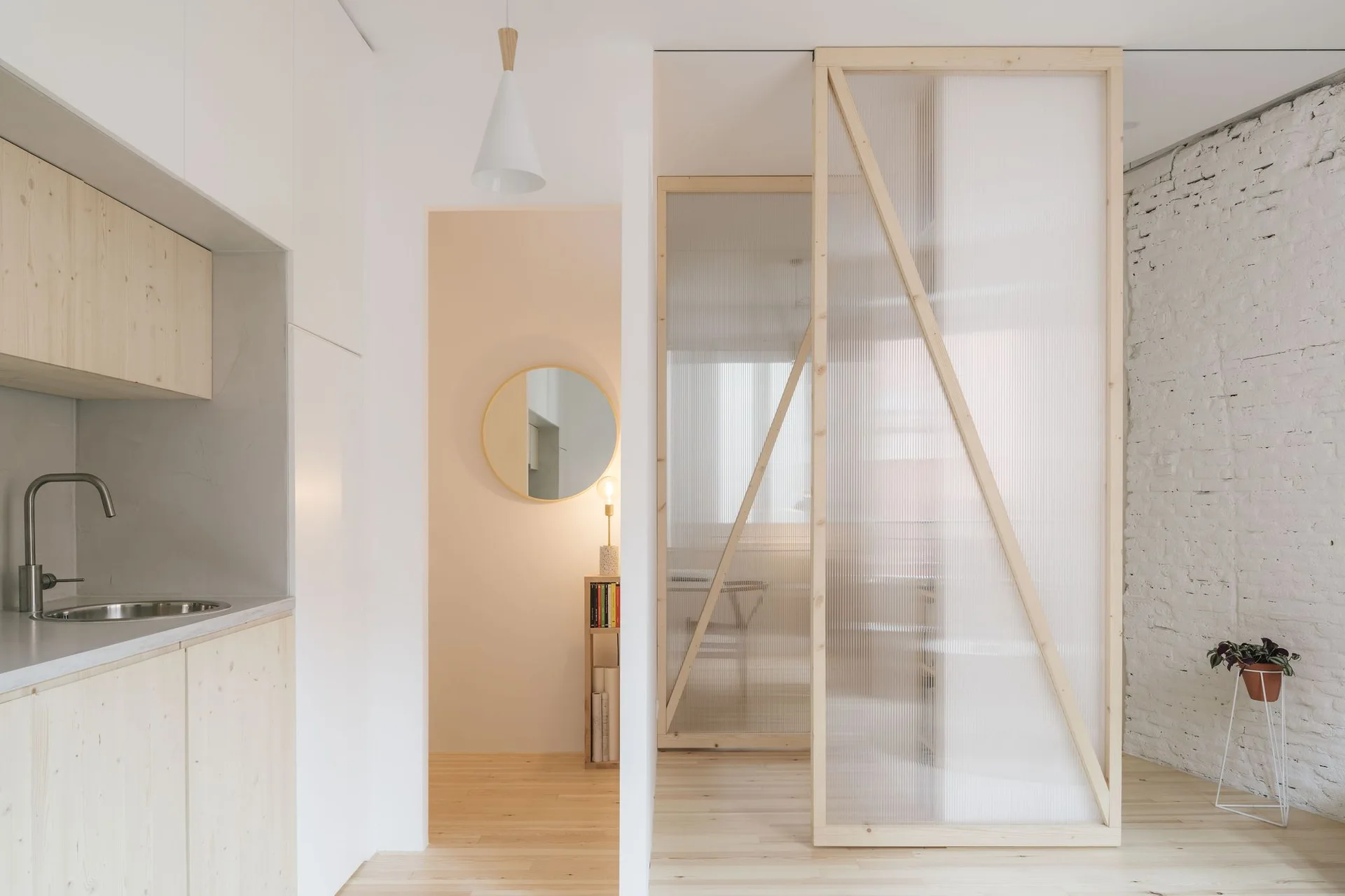 A bright minimalist kitchen with light wood cabinets is separated from another room by a timber-framed fluted glass sliding partition.