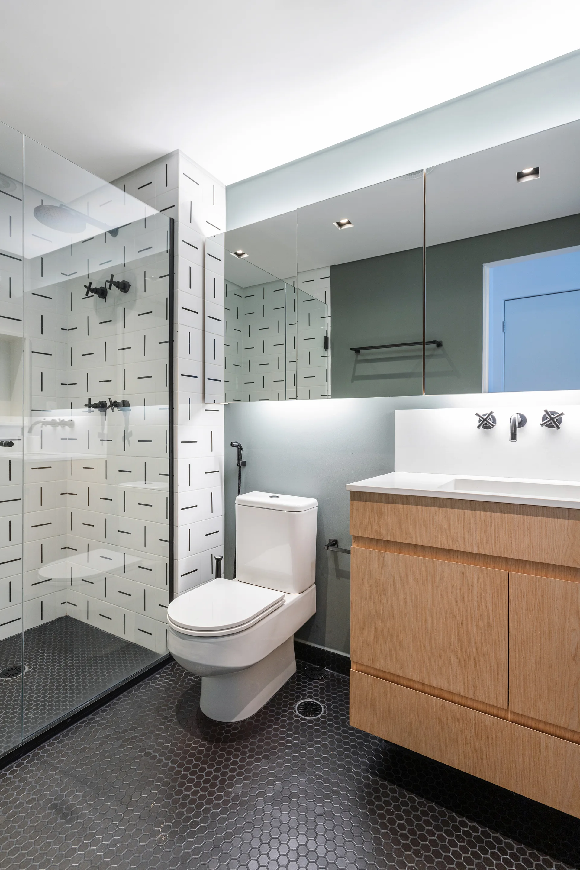 A modern bathroom featuring a floating wood vanity, a white toilet, and a glass-enclosed shower with graphic patterned wall tiles and black hexagonal flooring.