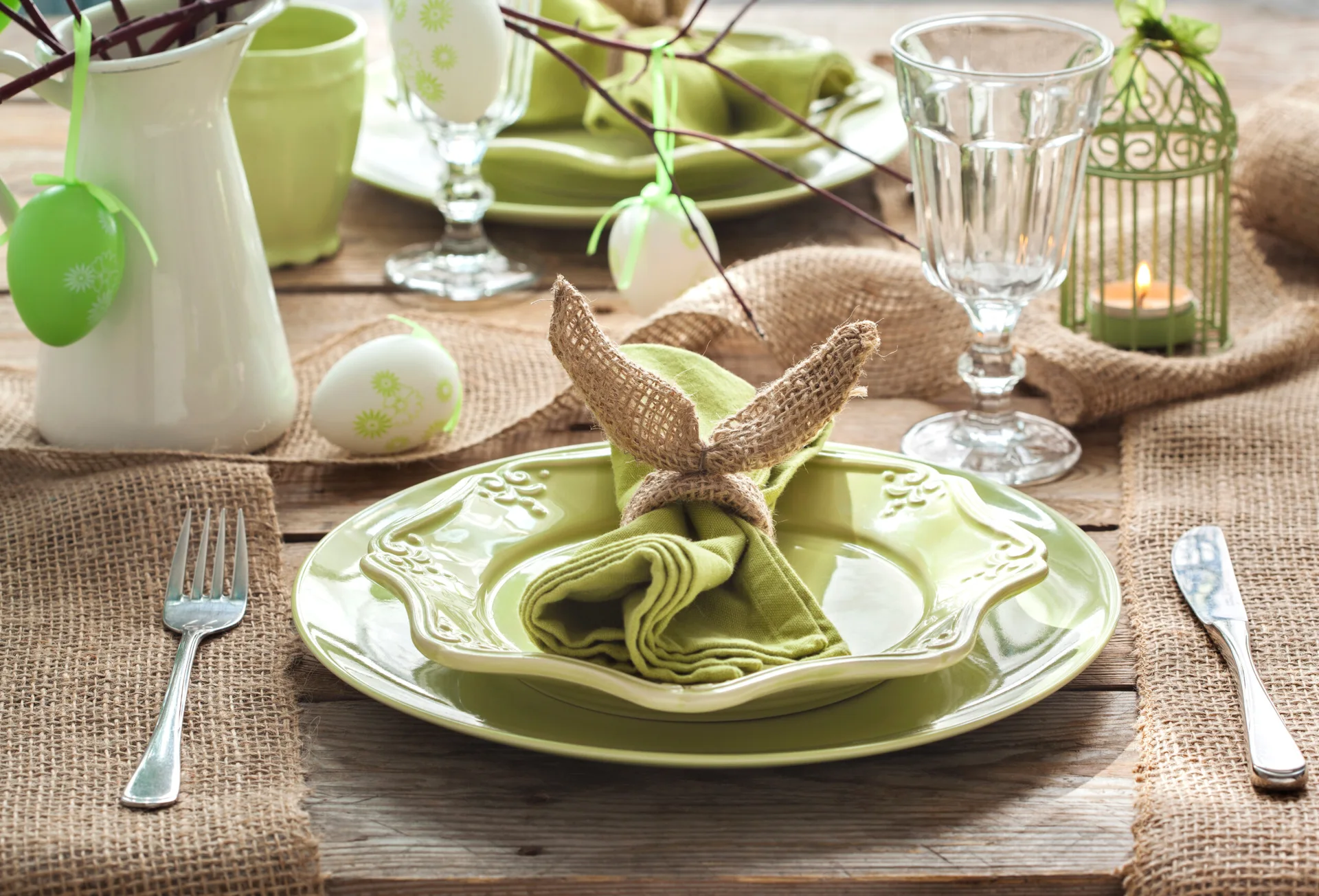 A rustic Easter table setting features sage green ceramic plates, a burlap runner, and a bunny-ear napkin ring on a weathered wooden tabletop.