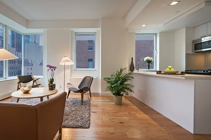 An open plan apartment living room features mid-century shell chairs, a leather armchair, a round wood coffee table, and a white kitchen counter.