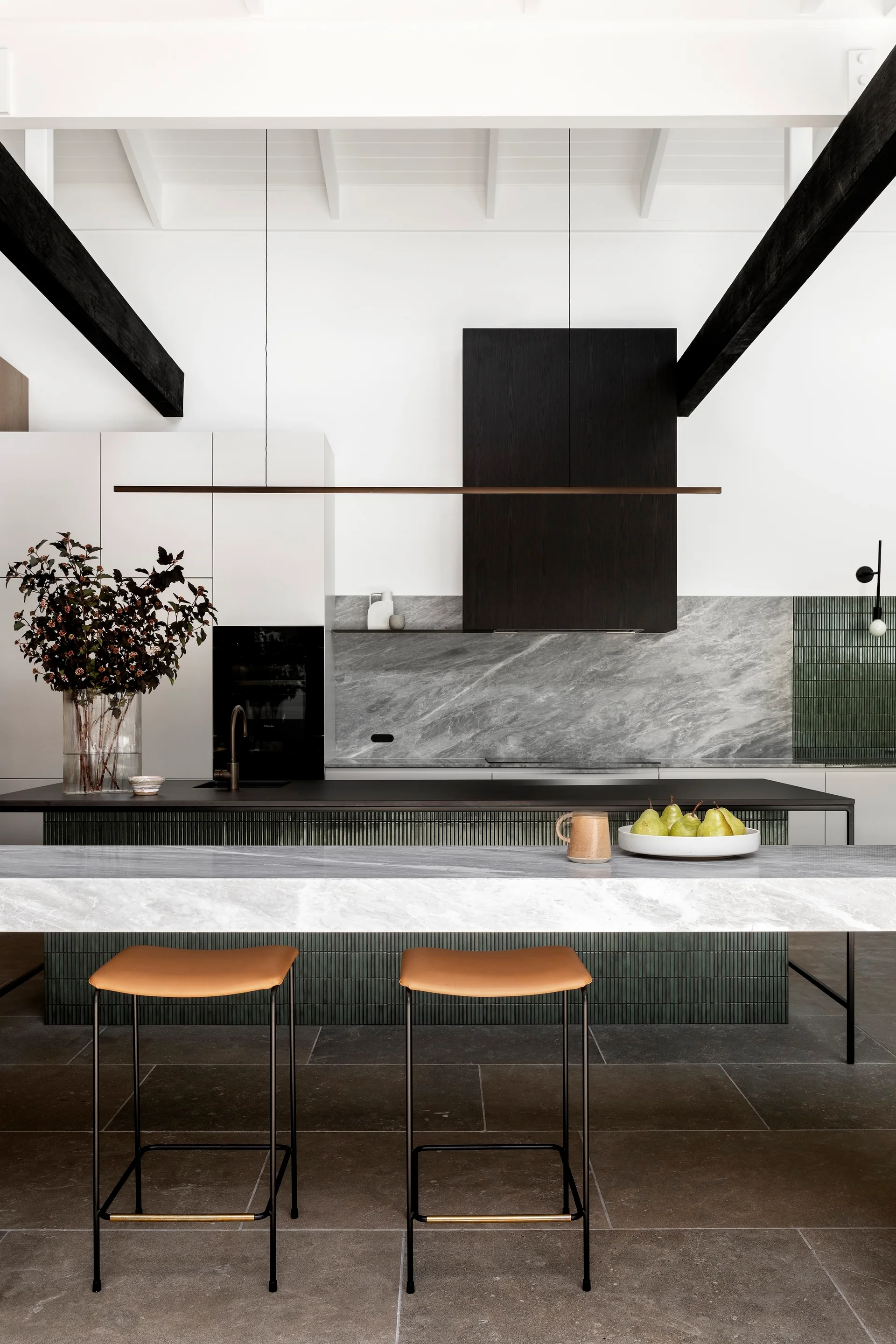 A modern kitchen features a marble-topped island with tan stools, dark ceiling beams, green ribbed tile cladding, and black cabinetry.