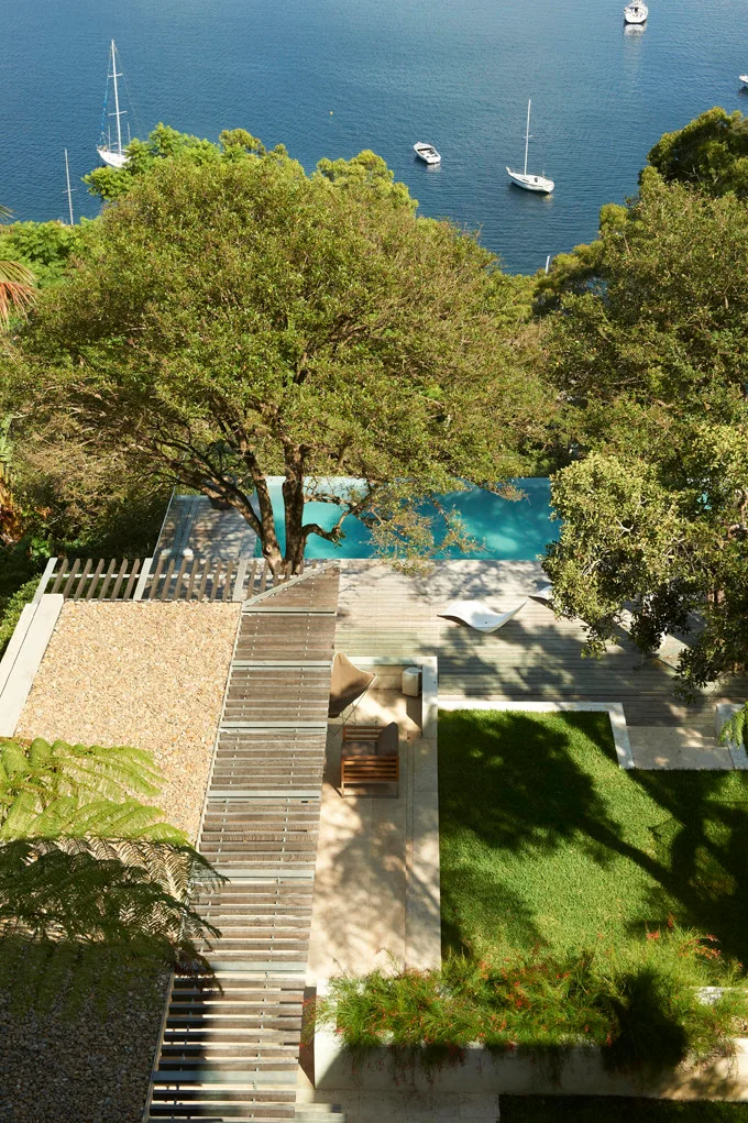 An overhead view of a terraced outdoor patio featuring a wooden deck, a bright blue swimming pool, a green lawn, and a view of the ocean.