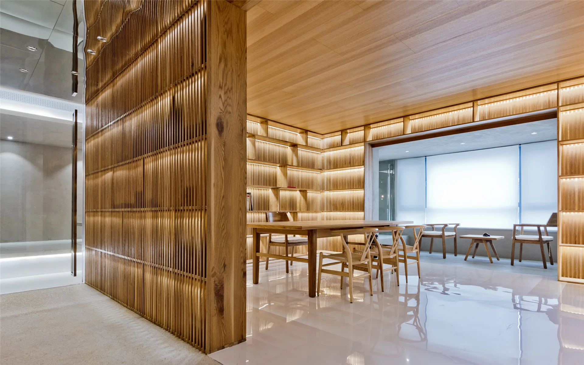 A minimalist dining room features a wooden dining table and chairs, integrated wood slat shelving with LED lighting, and a glossy white floor.