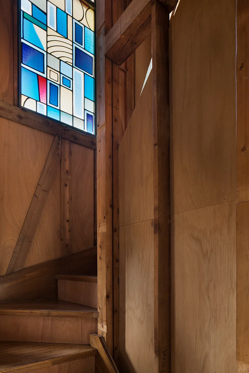 A wooden stairwell with warm plywood paneling and an overhead geometric stained glass window in shades of blue.