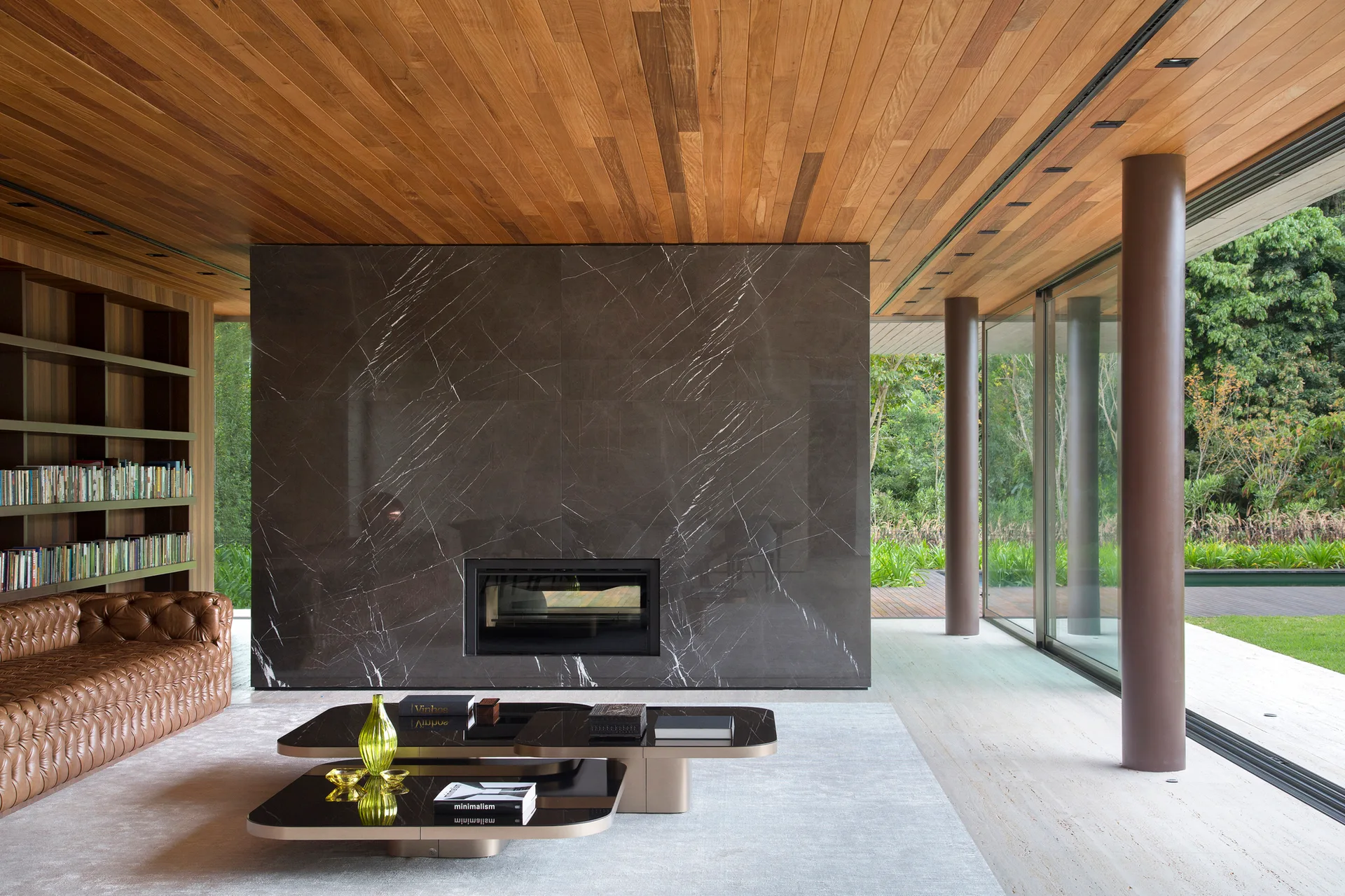 A modern living room with a wood plank ceiling features a tufted leather sofa, a dark marble fireplace divider, and floor-to-ceiling glass doors.