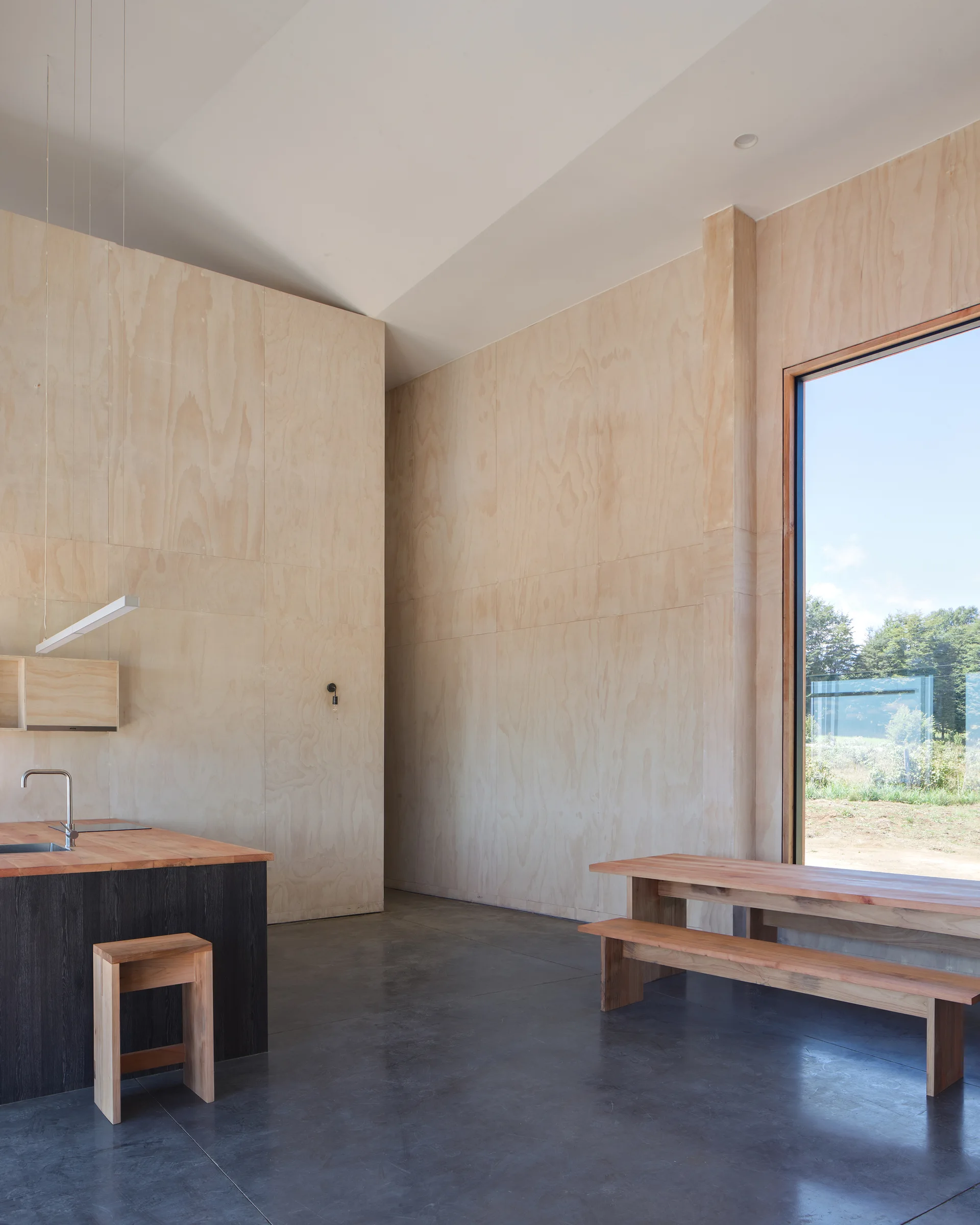 A minimalist kitchen and dining area with light plywood-paneled walls, a dark wood island, a wooden bench table, and polished concrete floors.