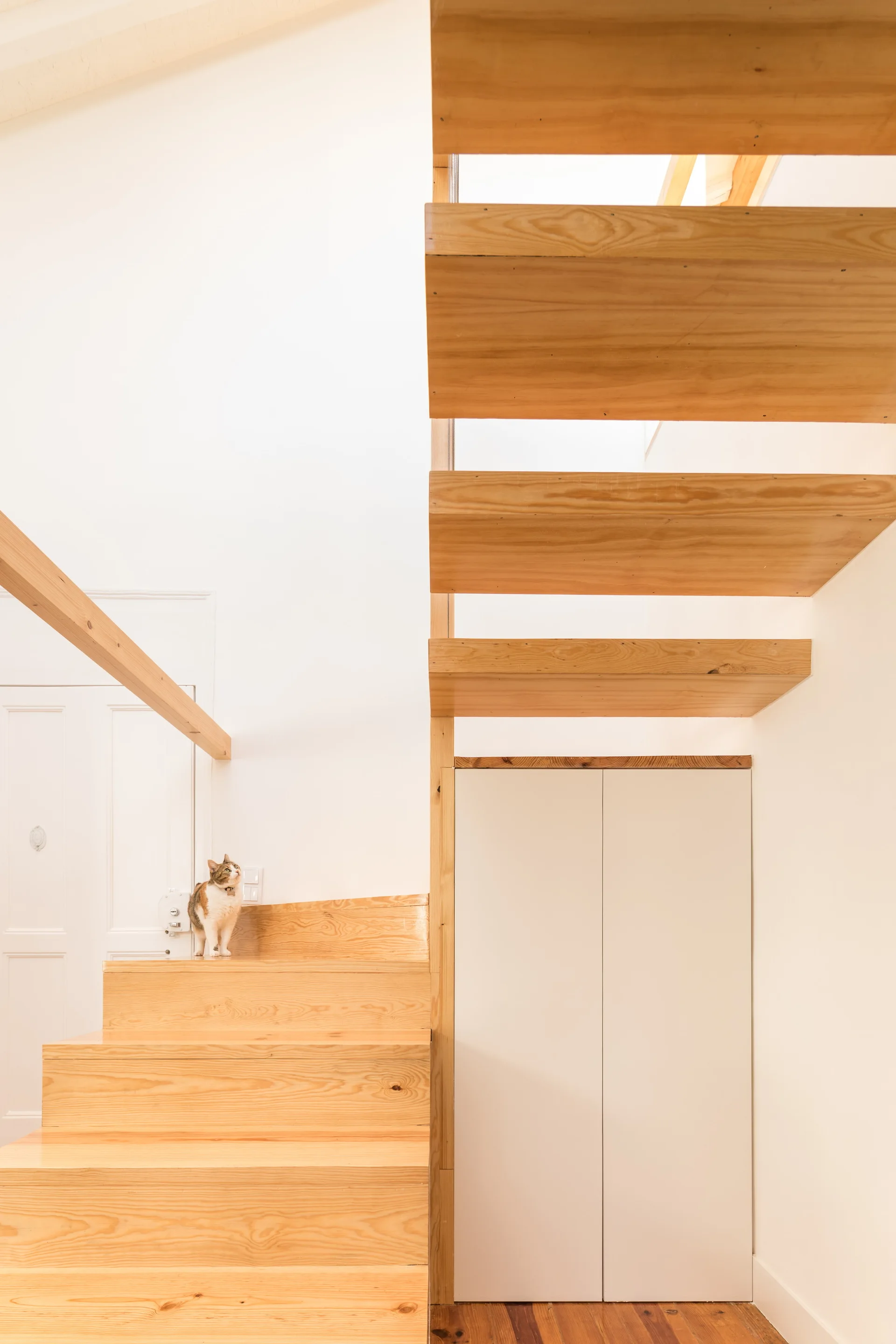 A minimalist floating wood staircase with open treads is positioned against a white wall next to a seamless built-in cabinet.